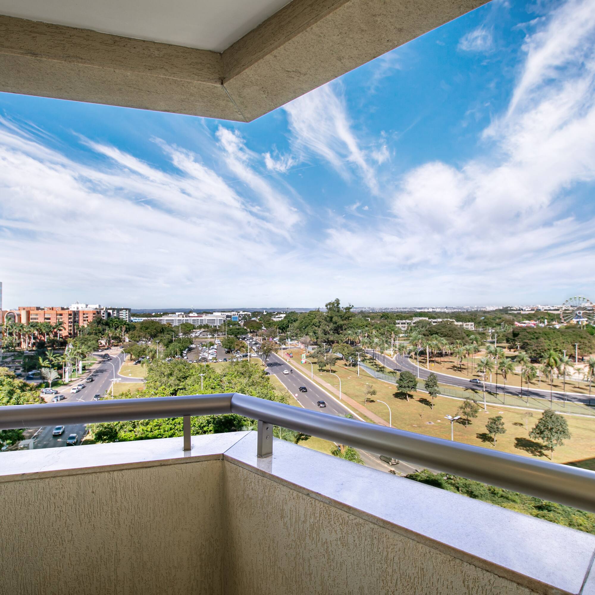 Balcony view: modern buildings, urban streets, green trees, and bright blue sky.
