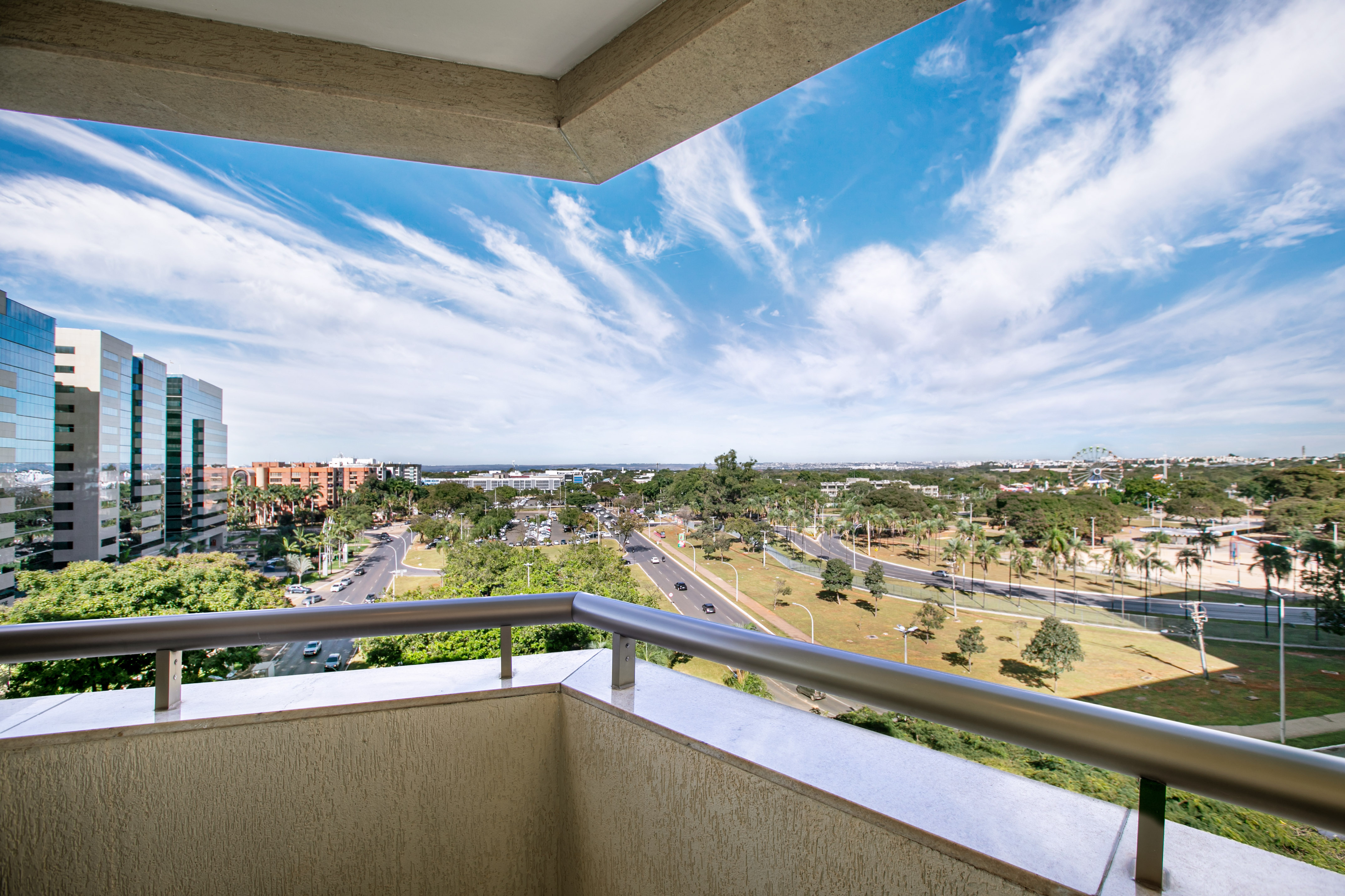 Balcony view: modern buildings, urban streets, green trees, and bright blue sky.