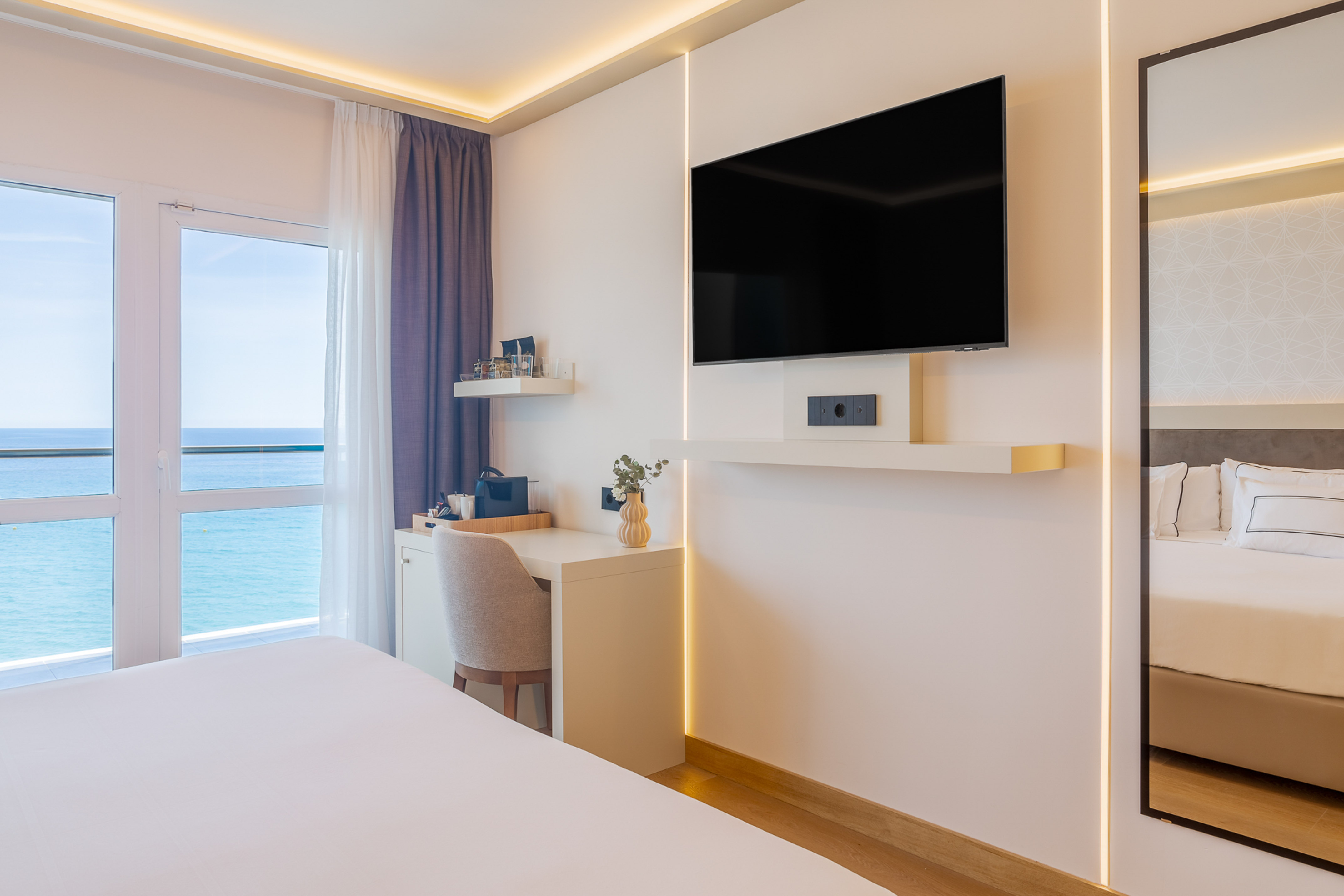 Bright hotel room with ocean view, white bed, flat-screen TV, and work desk.