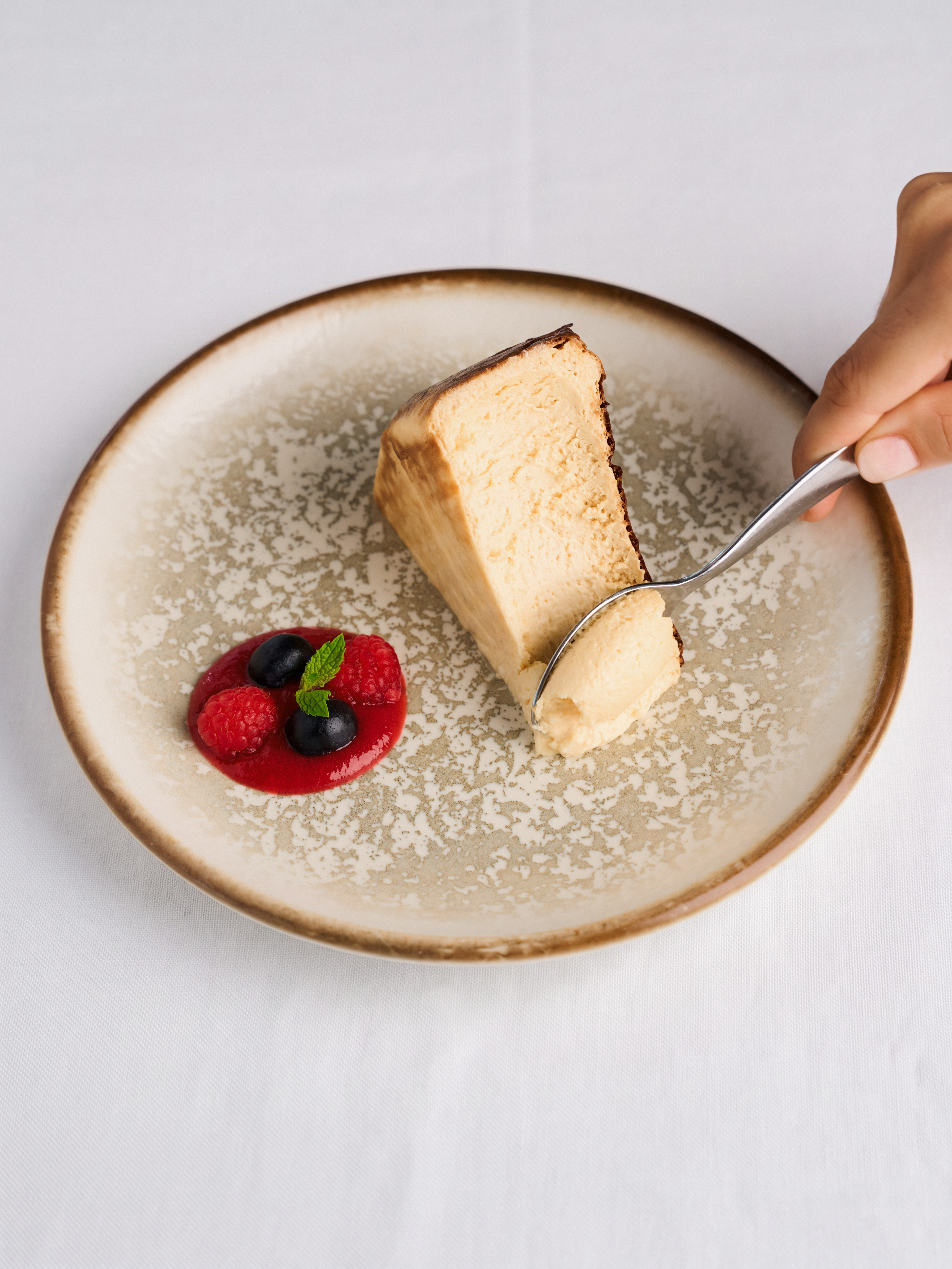 Cheesecake slice, berry sauce with raspberries & blueberries. Spoon lifting a bite.