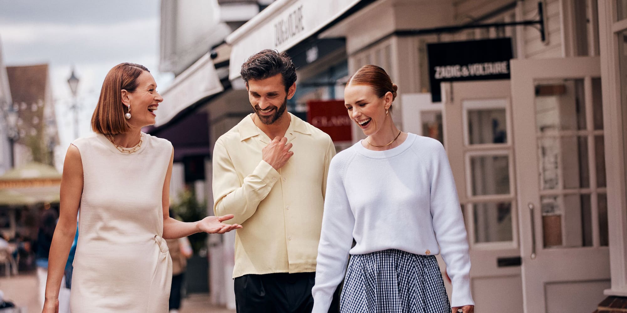 Three happy friends shopping in a luxury outlet village, passing Zadig & Voltaire.