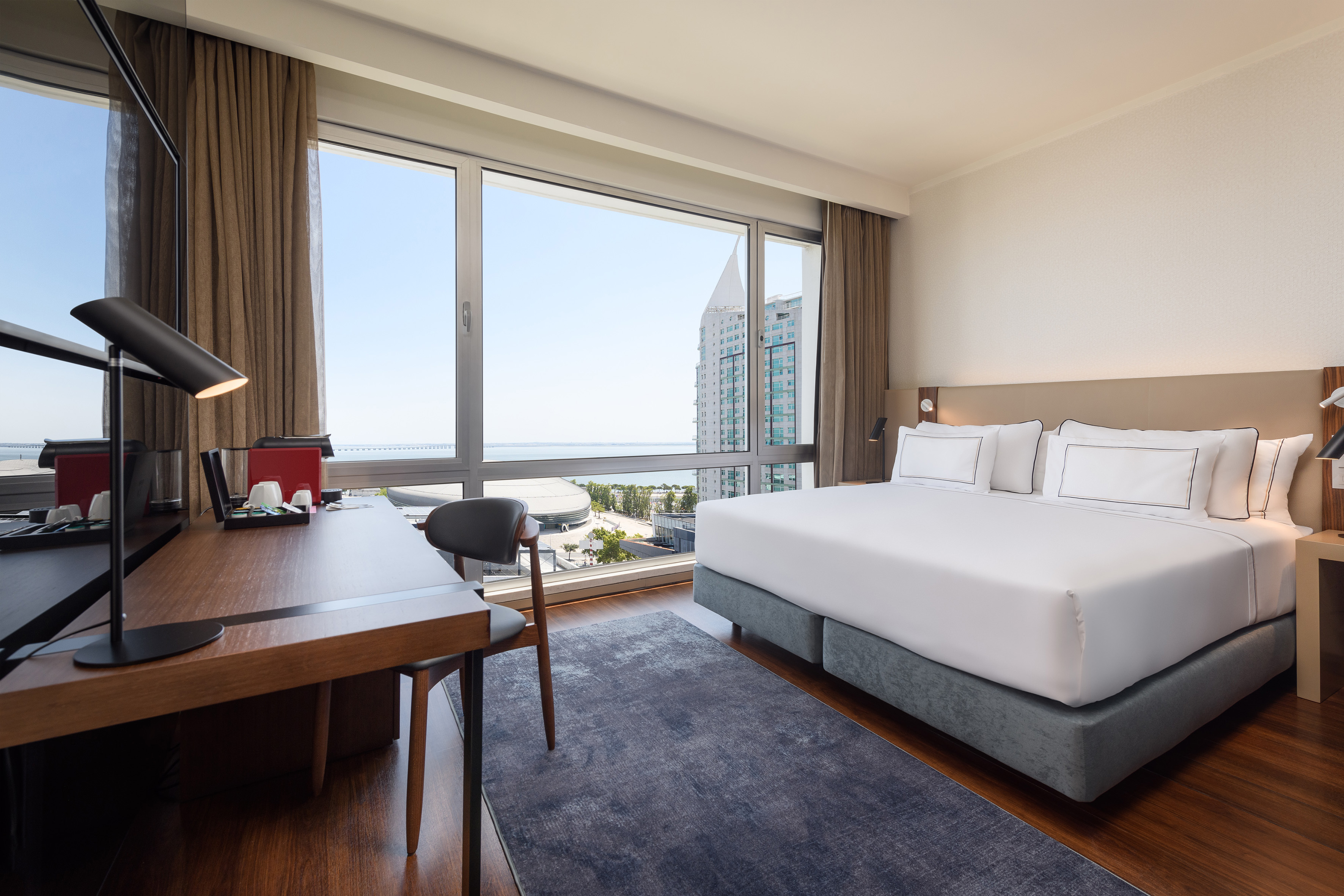 Modern hotel room with king bed, desk, and panoramic city water view.