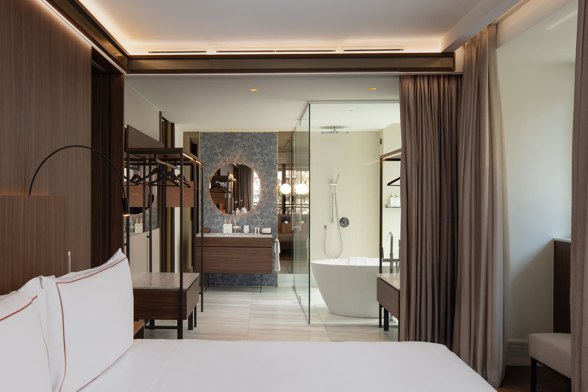 Modern hotel bedroom and open bathroom with freestanding tub, vanity.