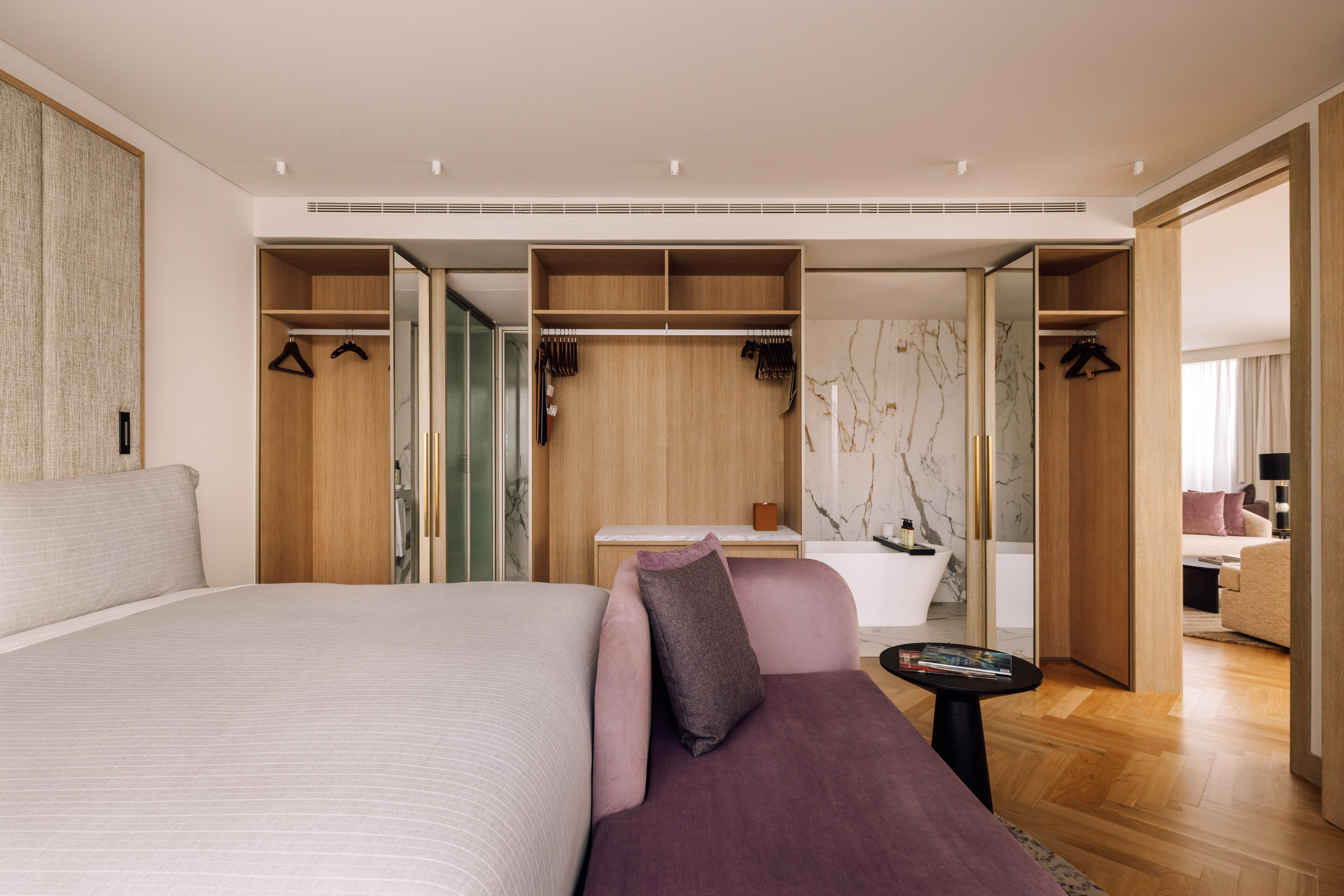 Modern hotel suite with bed, purple chaise, wood wardrobe, and marble bath.