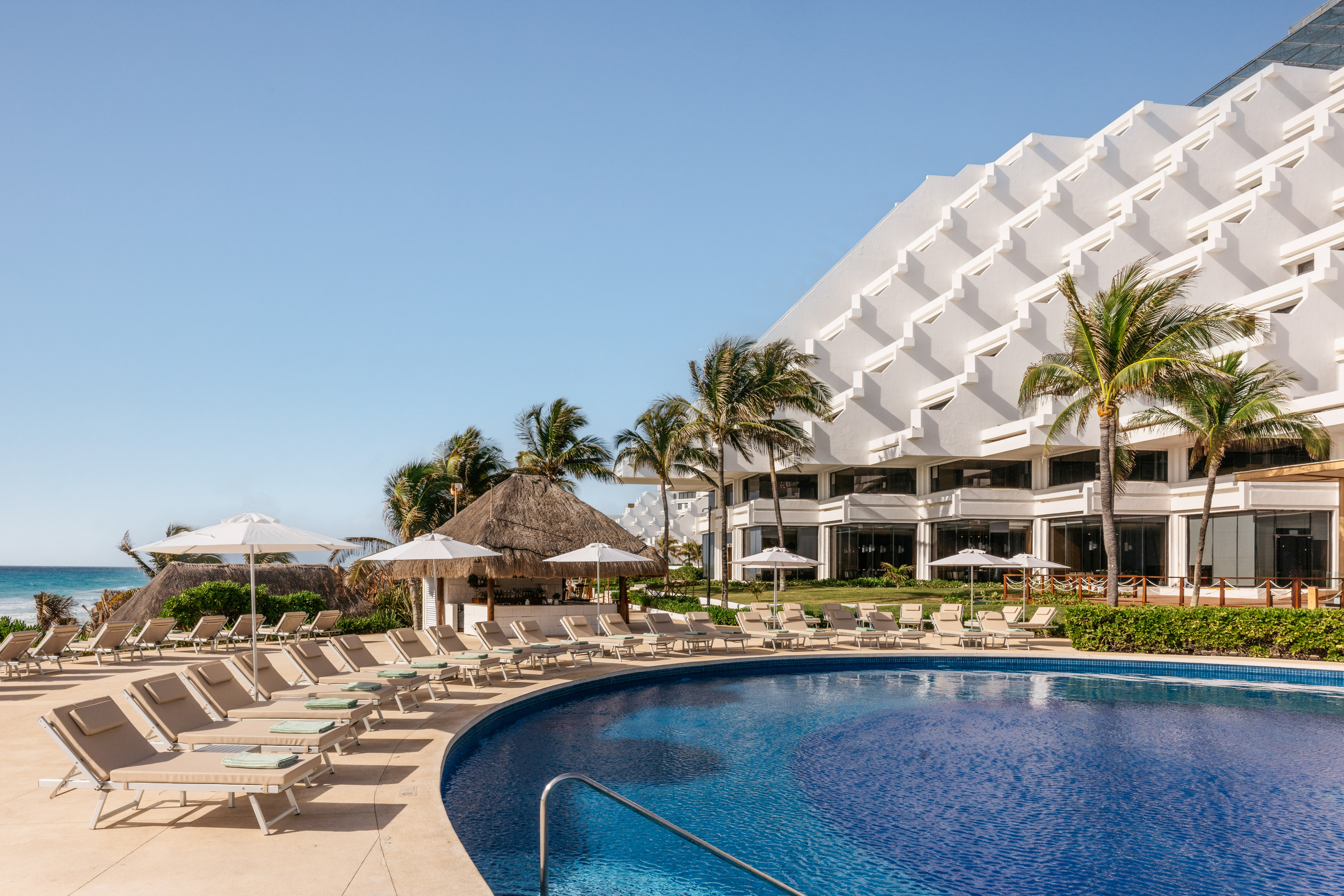 Resort pool with lounge chairs, palm trees, modern hotel, and ocean view.