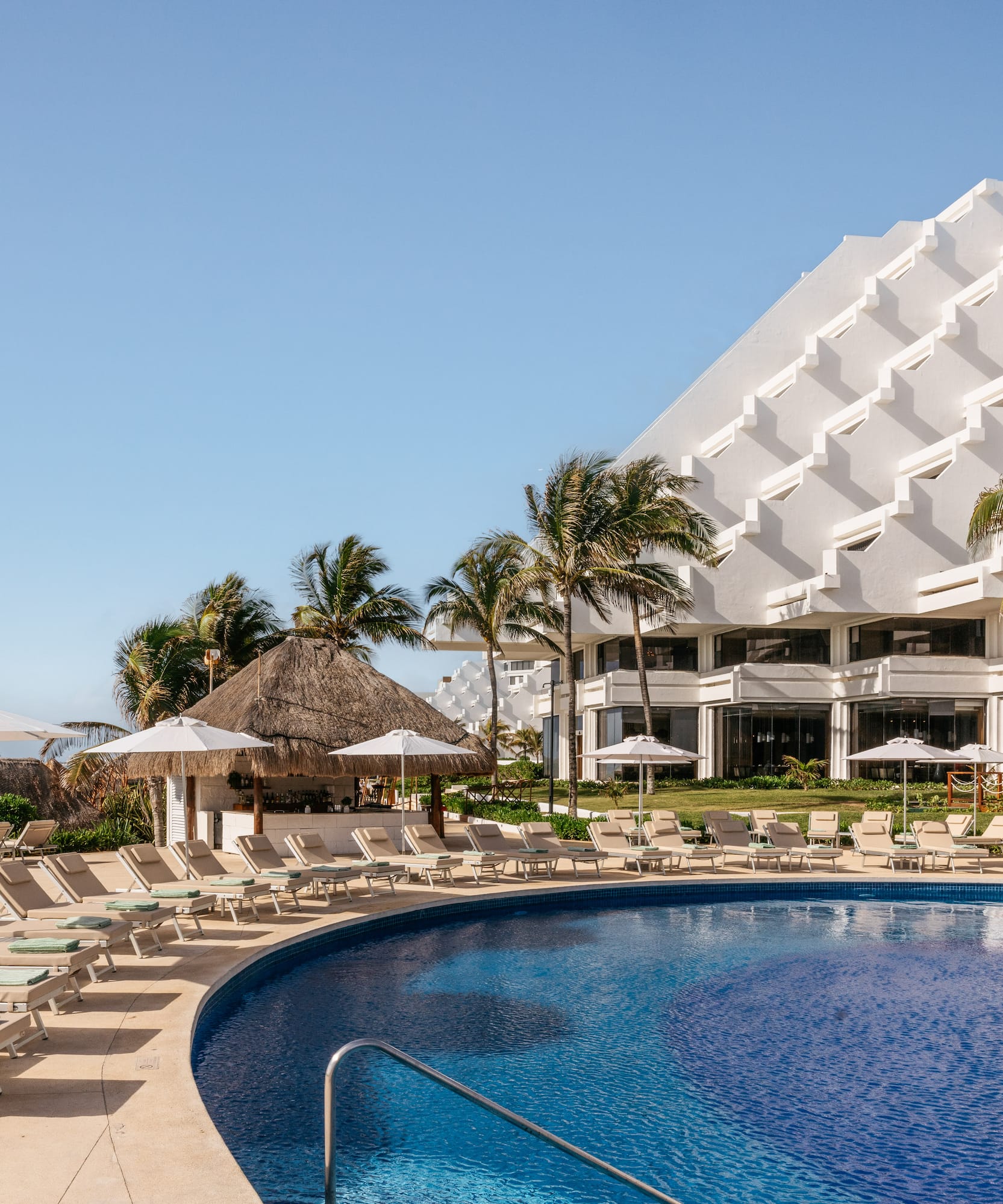 Resort pool with lounge chairs, palm trees, modern hotel, and ocean view.