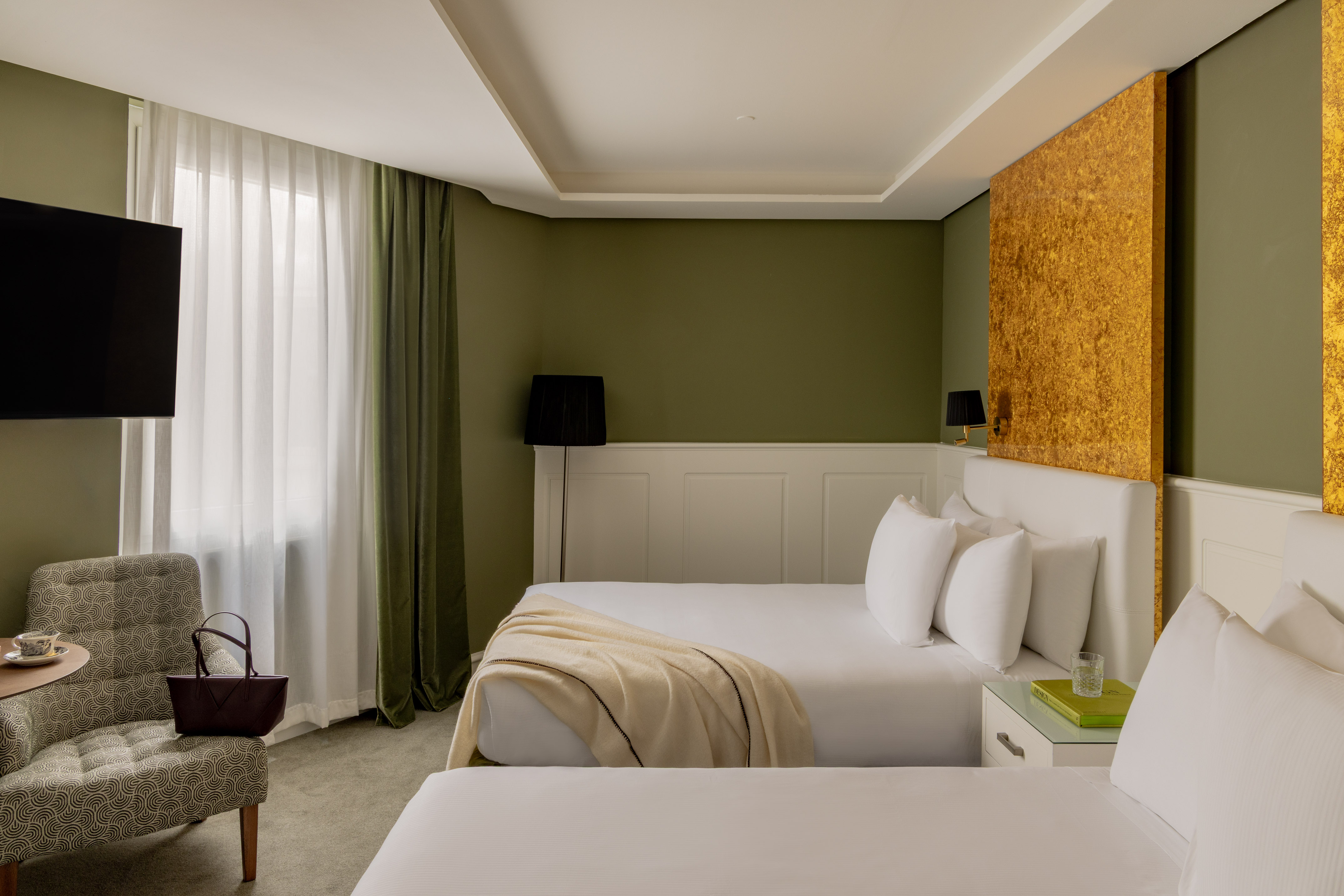 Modern hotel room with twin beds, olive green walls, and golden accent panels.