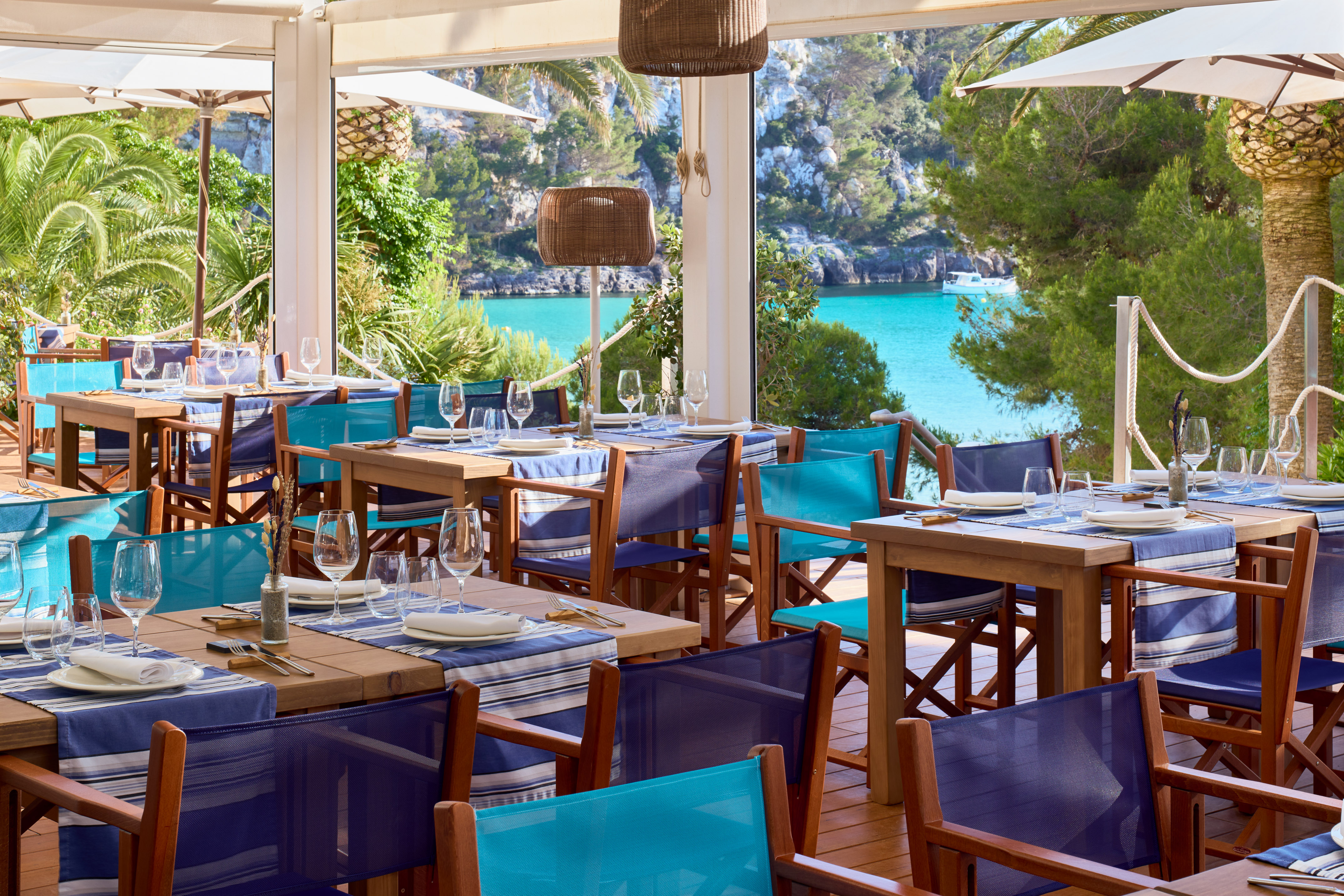 Coastal restaurant with blue chairs, set tables, and turquoise sea view.