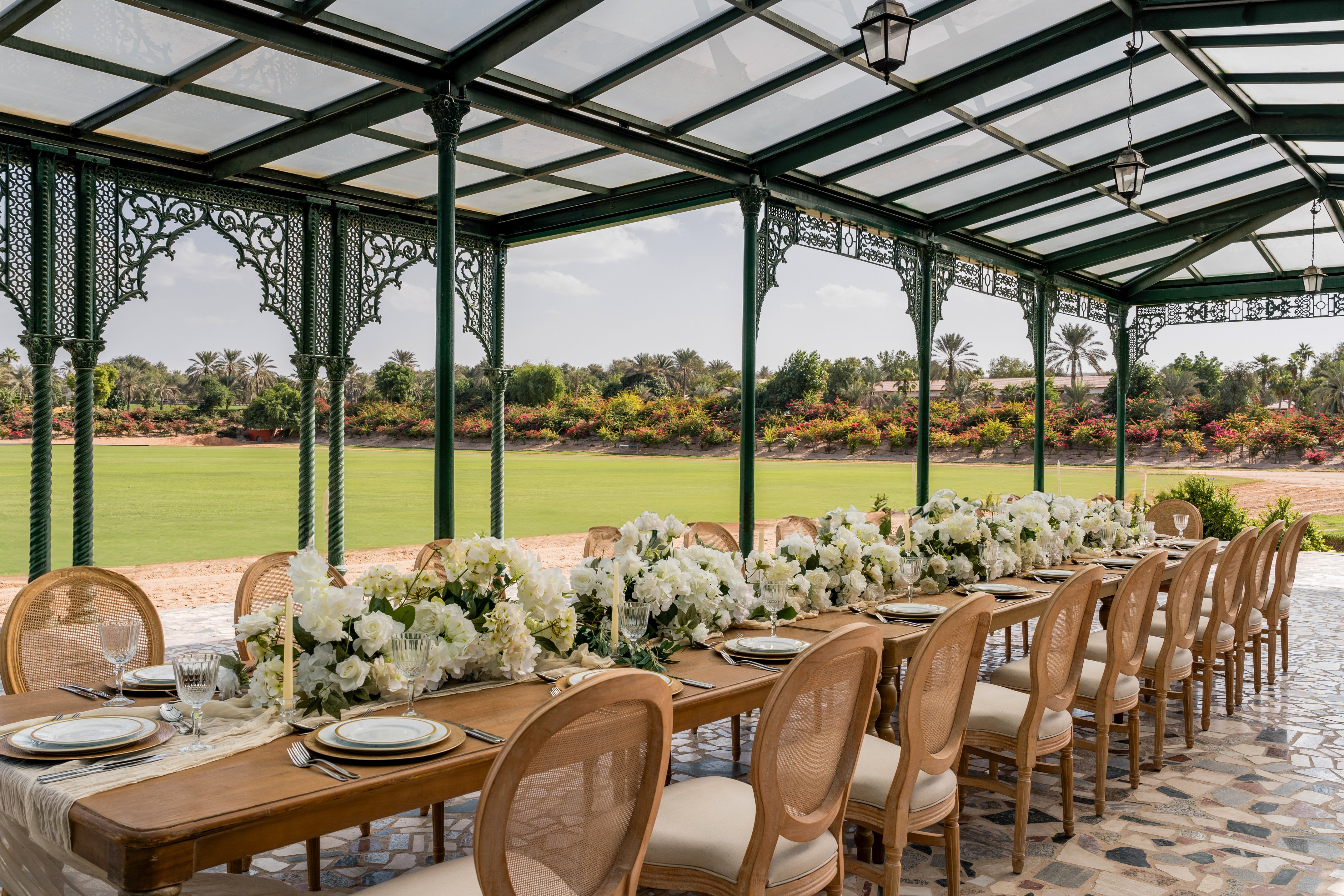 Outdoor dining setup with white florals under a gazebo, green polo field.