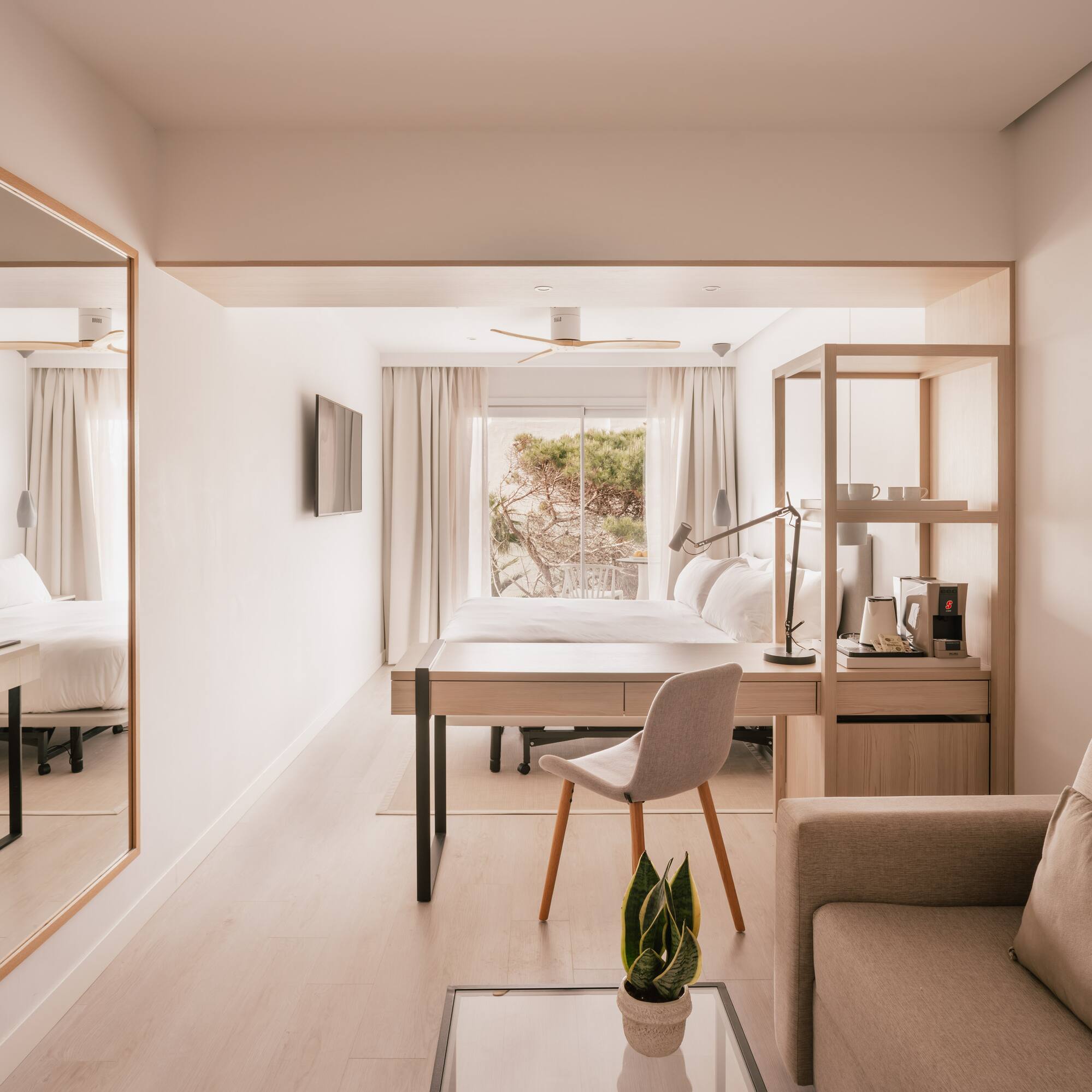 Modern light-filled hotel suite with bed, desk, sofa, and nature view.