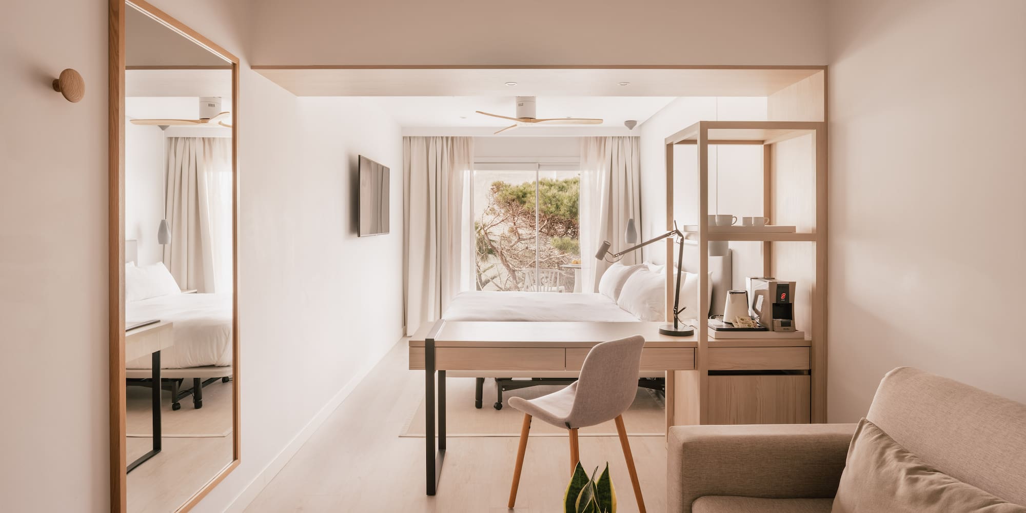 Modern light-filled hotel suite with bed, desk, sofa, and nature view.