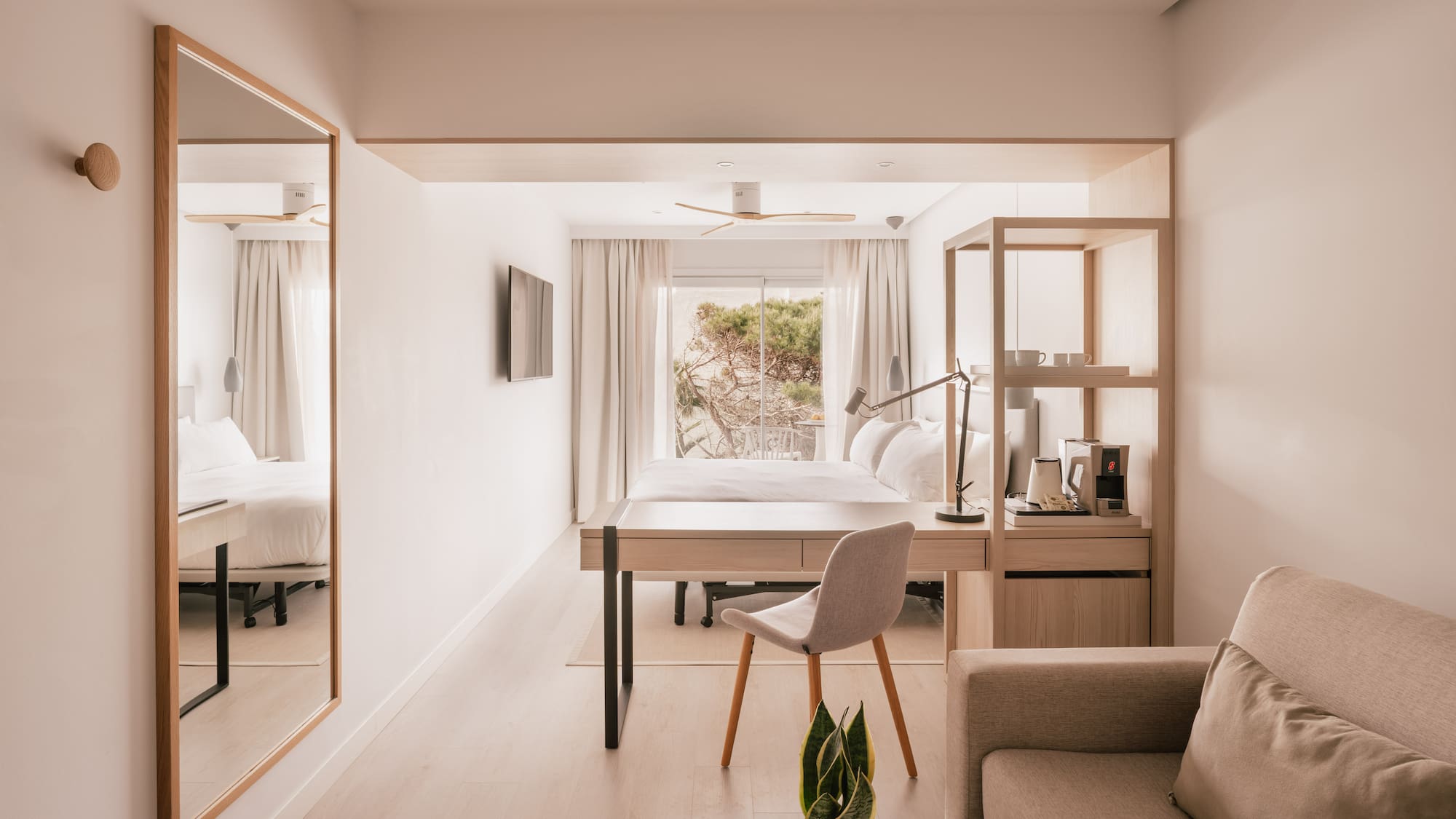 Modern light-filled hotel suite with bed, desk, sofa, and nature view.