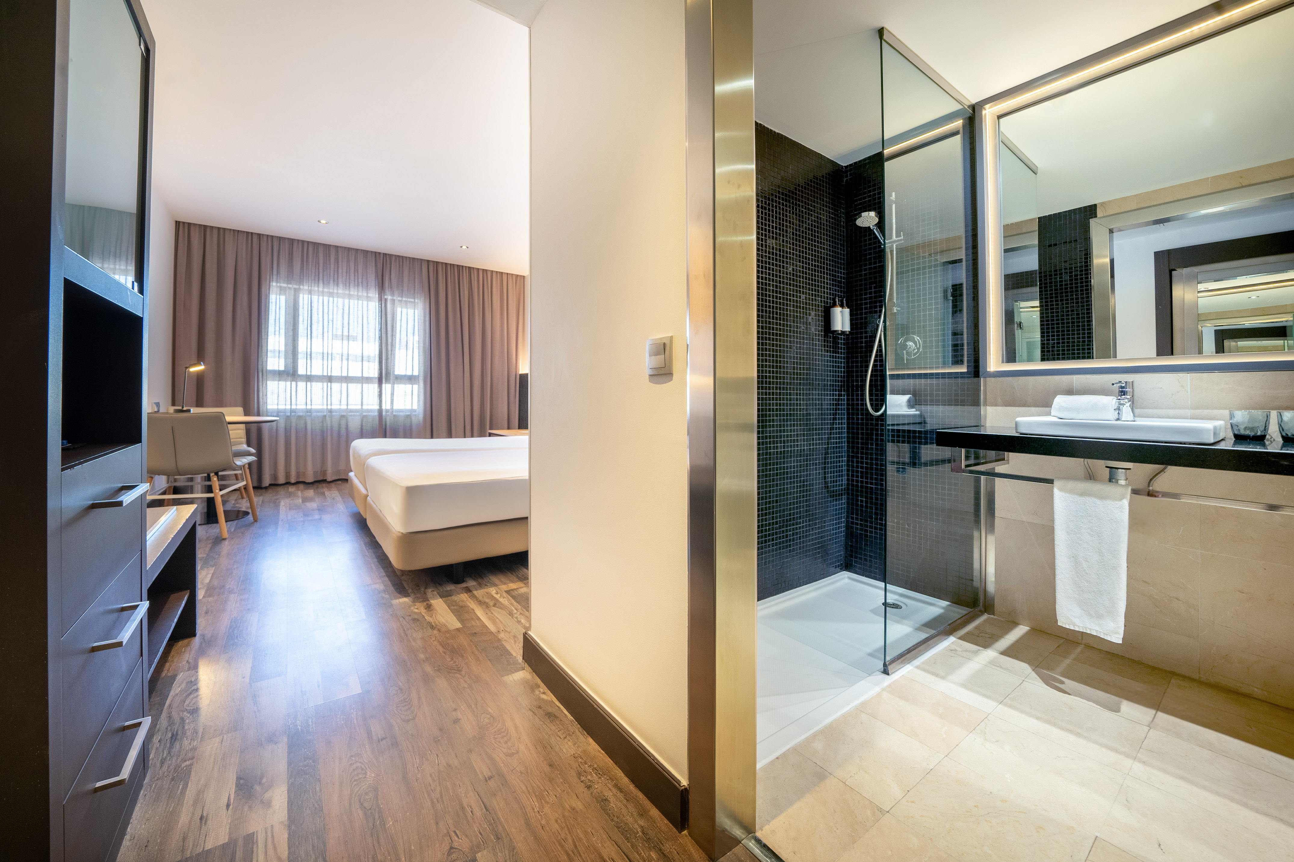 Modern hotel guestroom with twin beds, desk, and bathroom with glass shower.