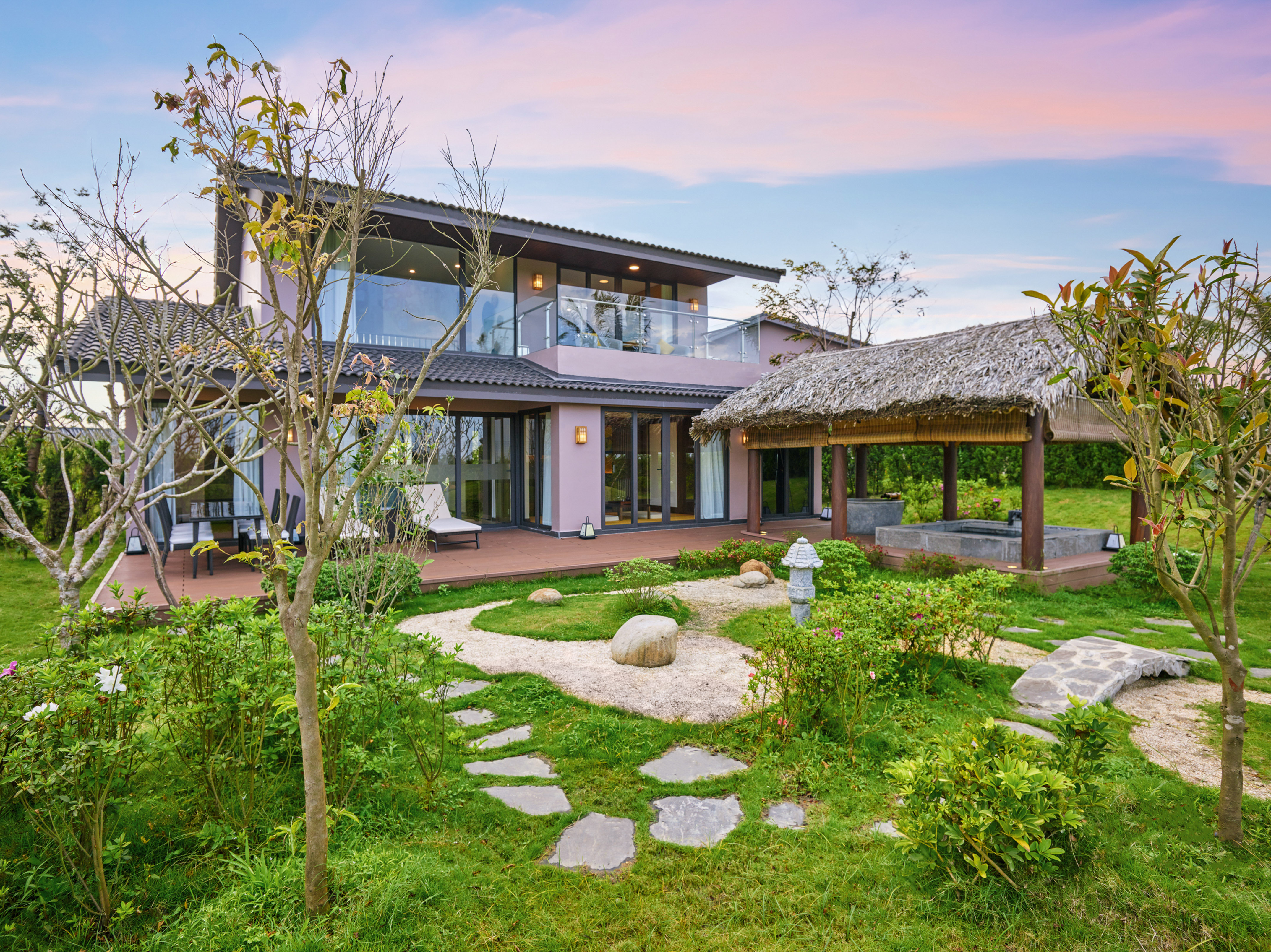 Modern villa with garden, thatched gazebo, hot tub, and stone path at sunset.