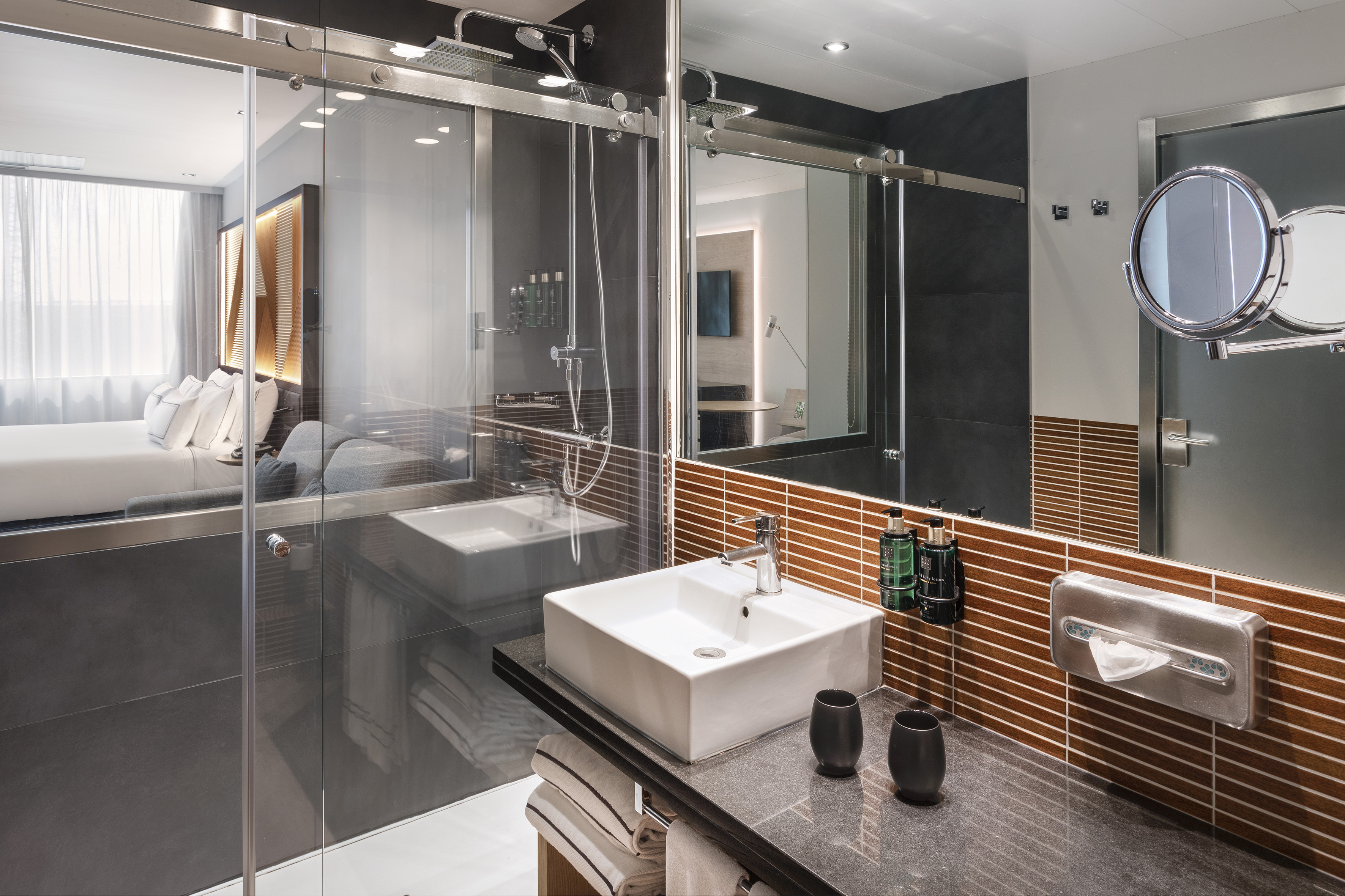 Modern hotel bathroom with white sink, glass shower, and bedroom view.