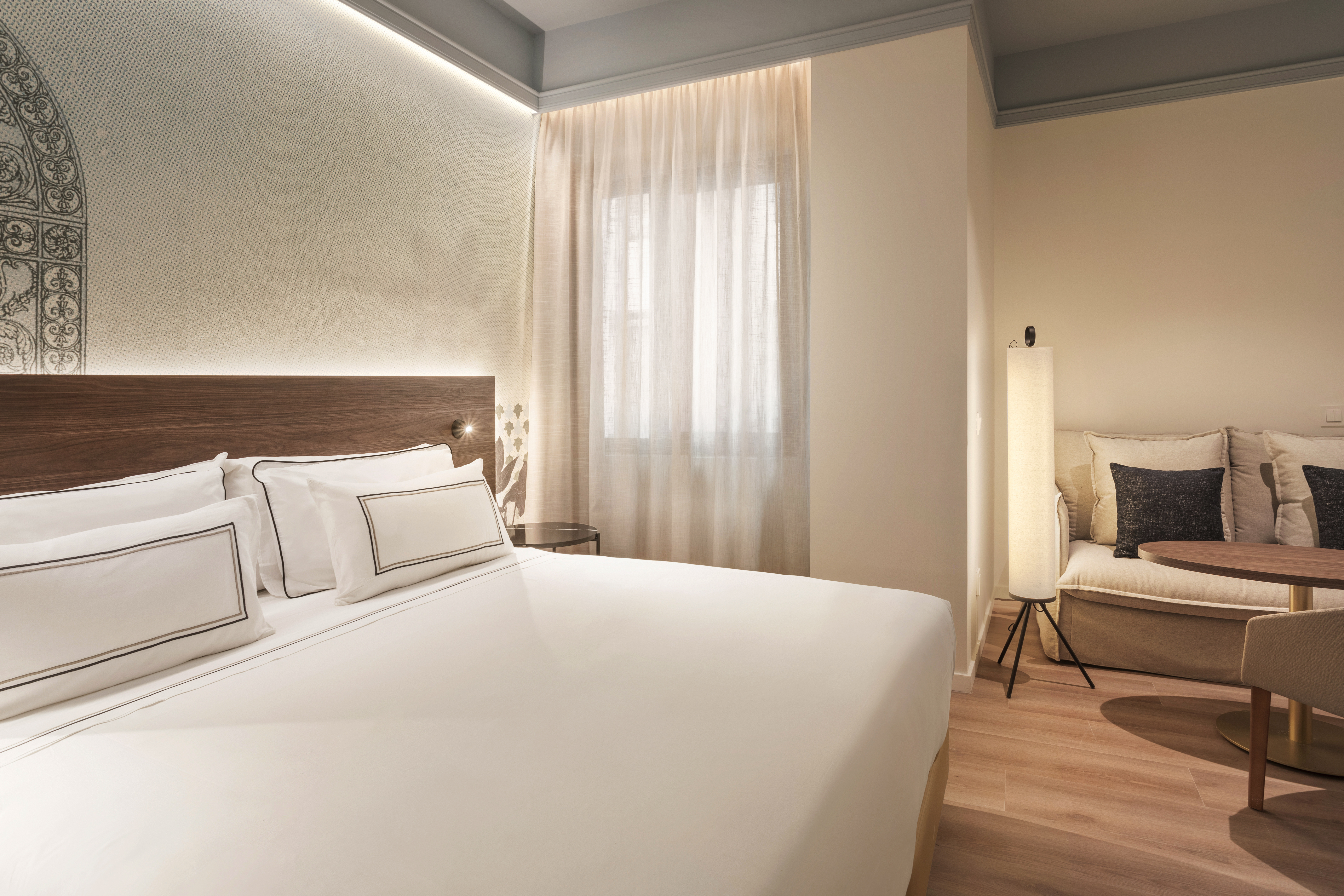 Modern hotel bedroom with large bed, wooden headboard, and cozy lounge area.