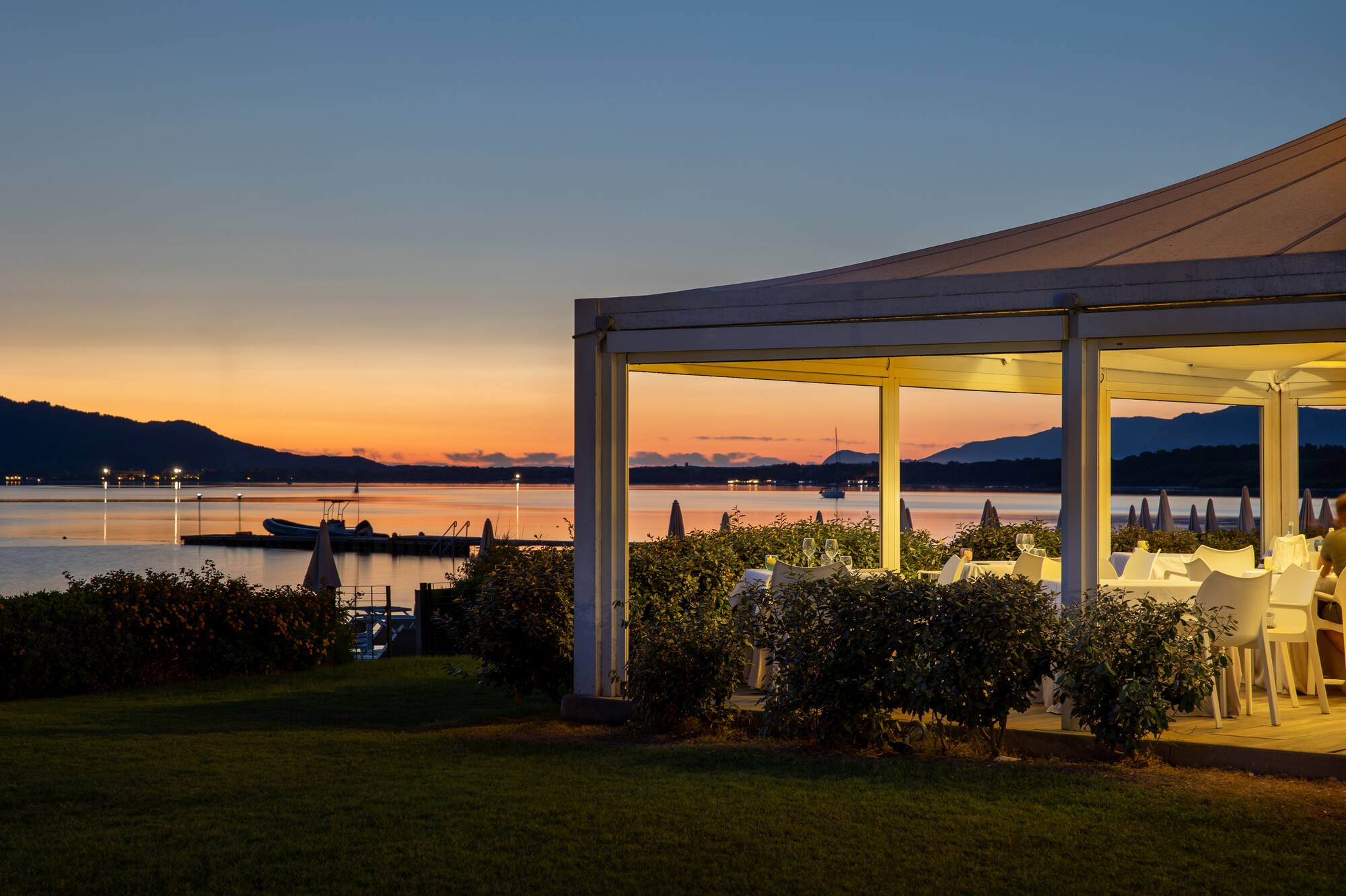 Illuminated outdoor restaurant by bay at sunset with mountains in background.