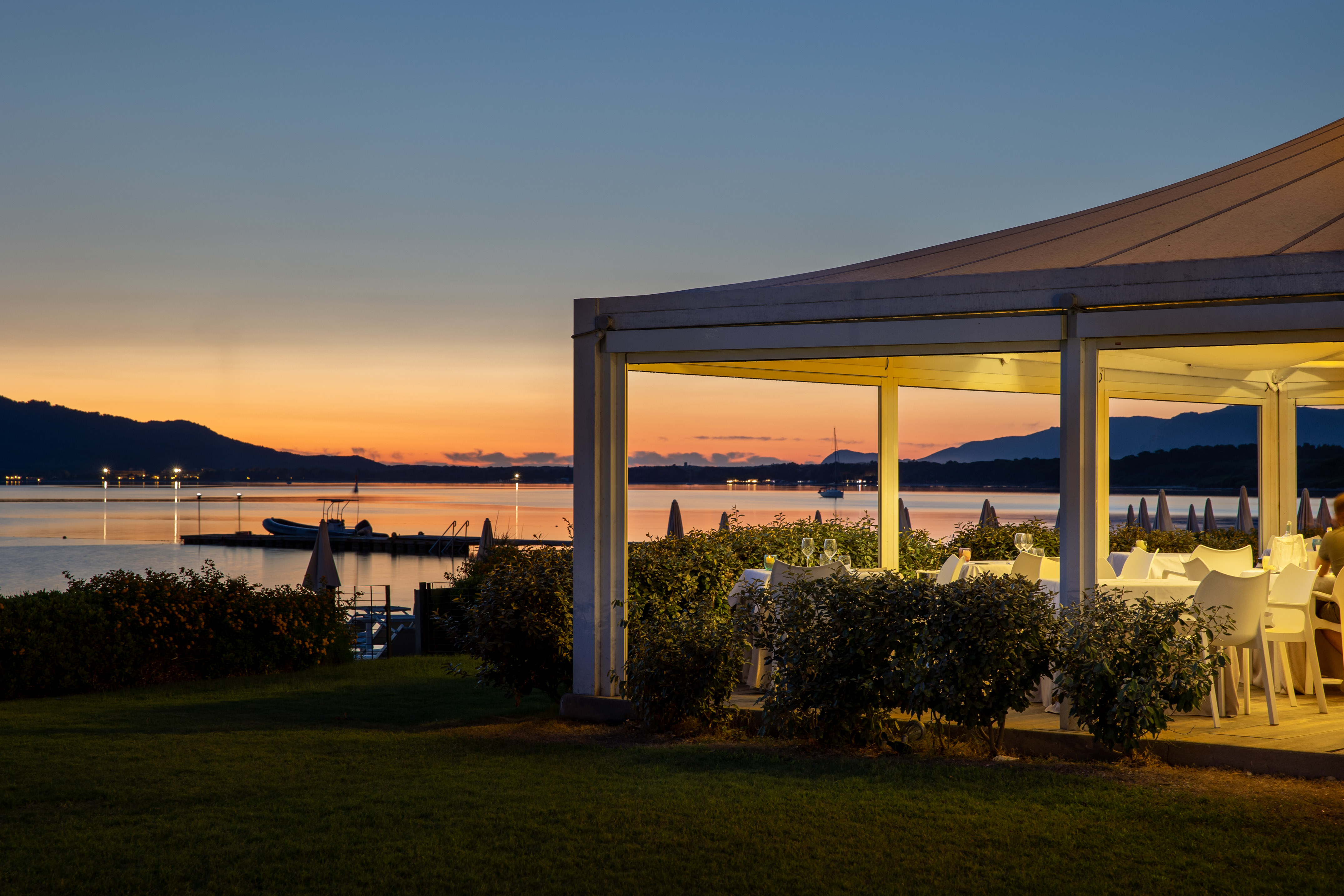 Illuminated outdoor restaurant by bay at sunset with mountains in background.