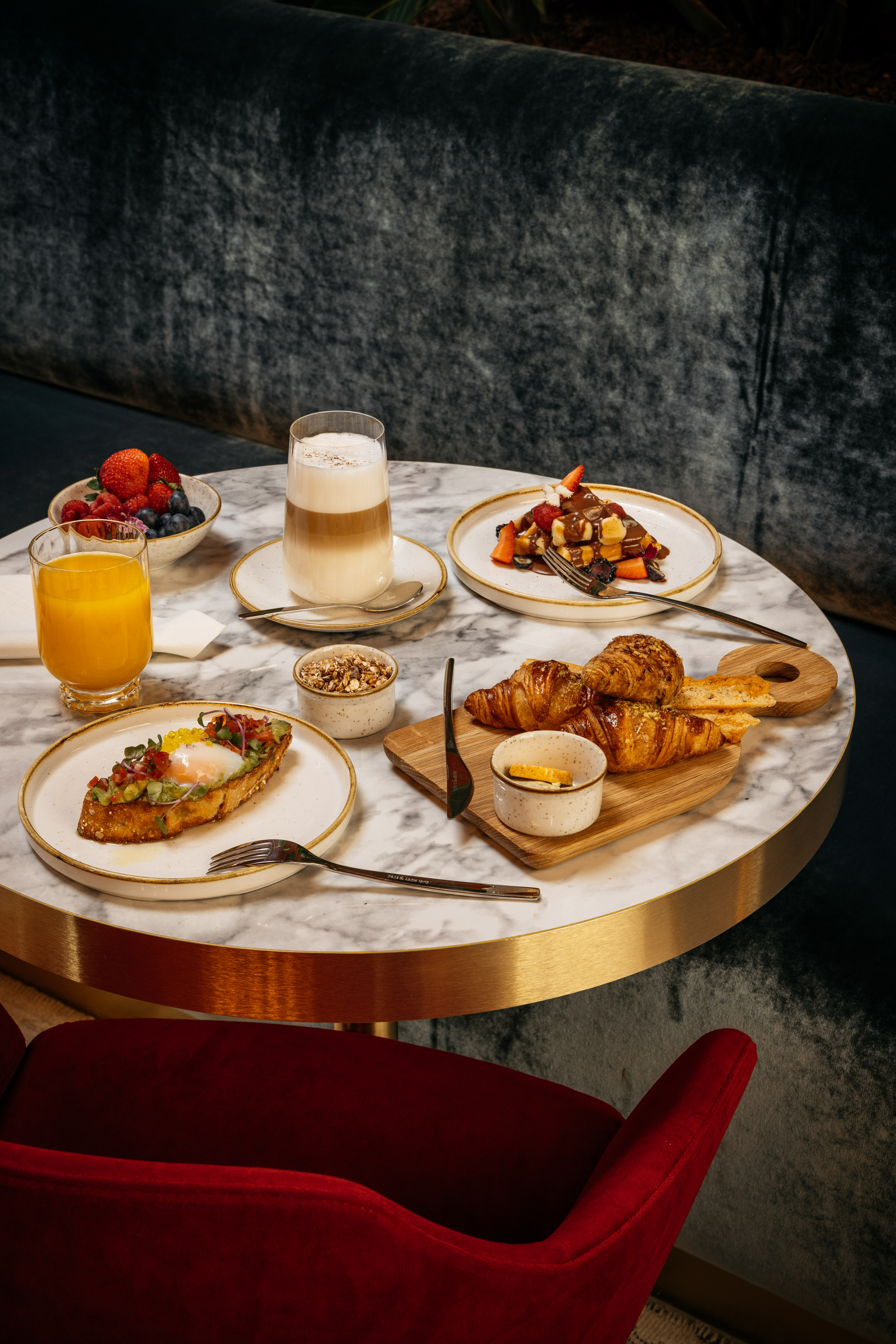 Brunch on marble table: avocado toast, waffles, croissants, berries, juice, latte.