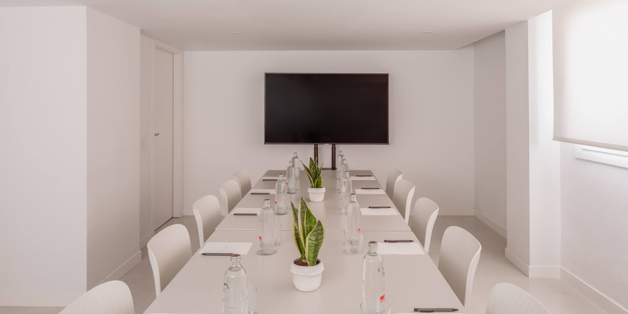 Moderna sala de reuniones blanca con pantalla, mesa de conferencias y plantas.
