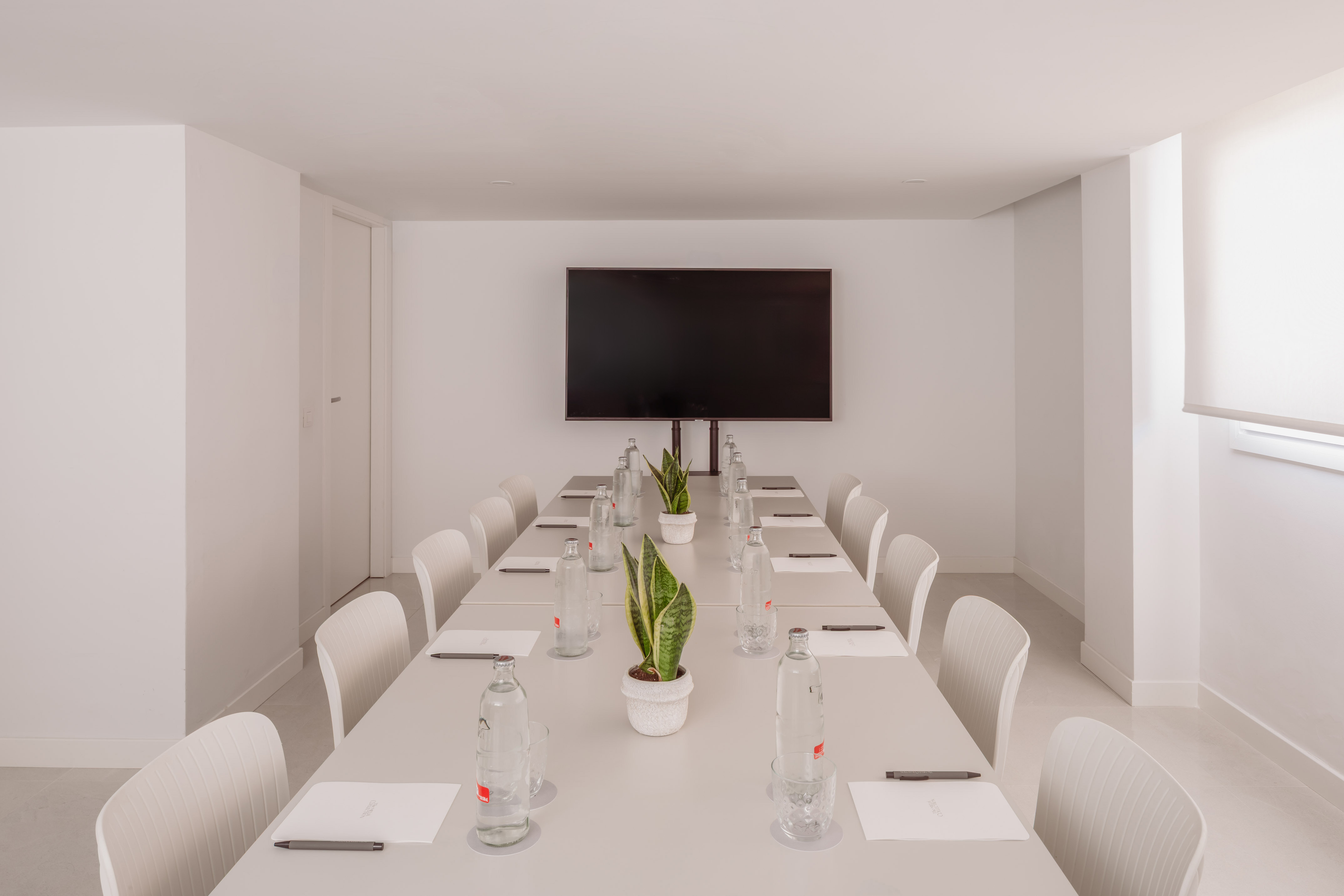Moderna sala de reuniones blanca con pantalla, mesa de conferencias y plantas.
