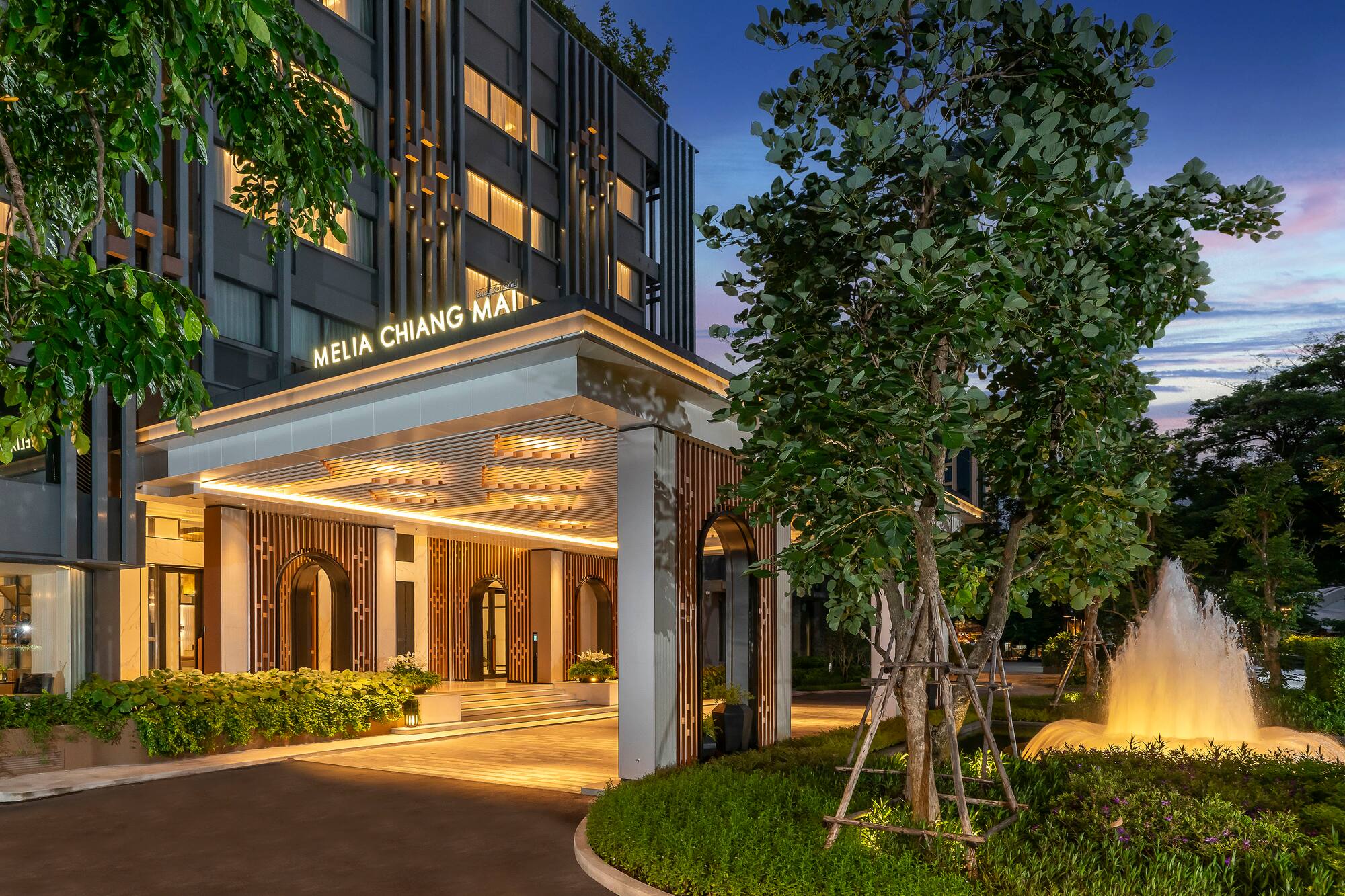Melia Chiang Mai hotel entrance at dusk, illuminated, with fountain and trees.