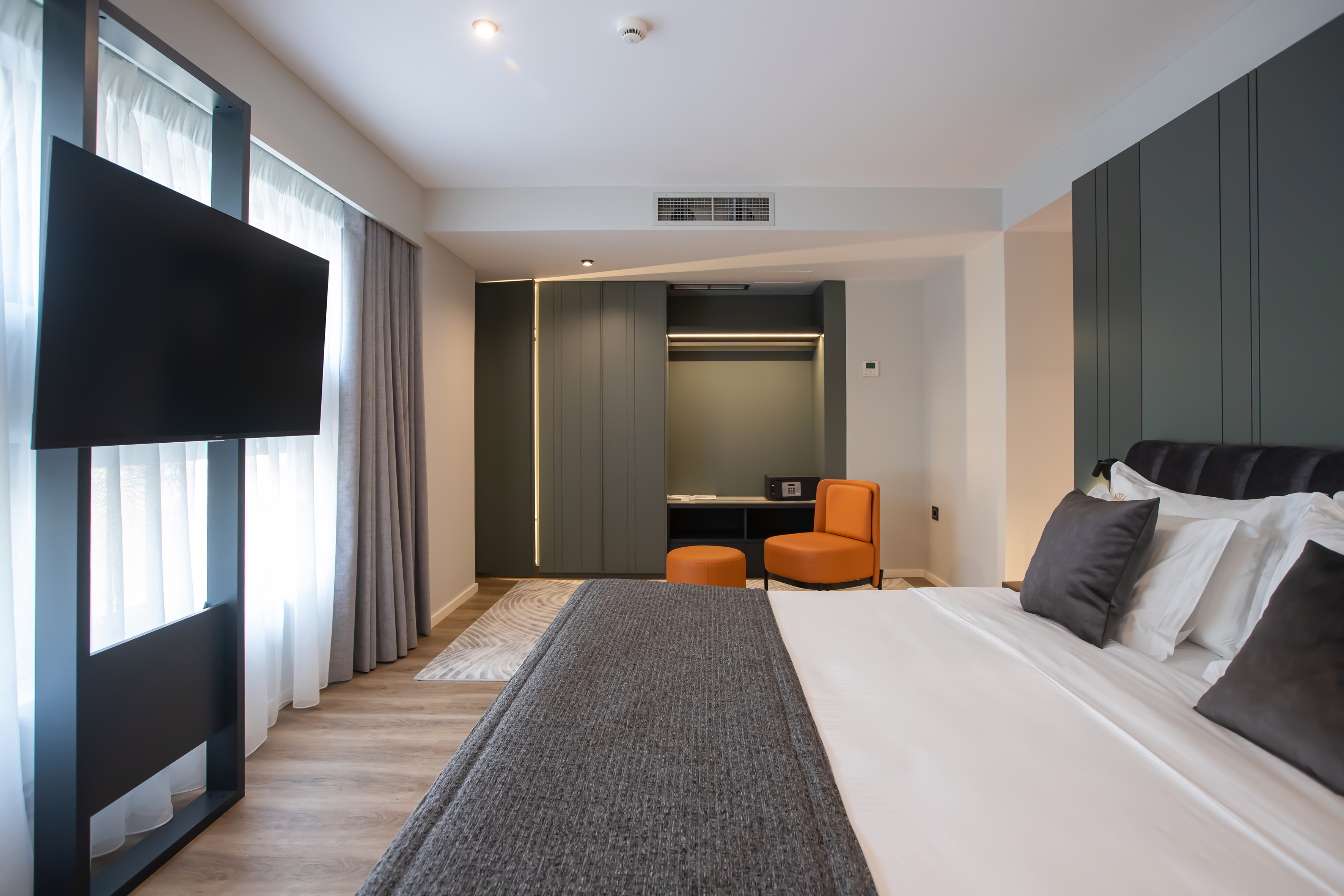 Modern hotel bedroom with large bed, orange accent chair, wardrobe, and wall TV.