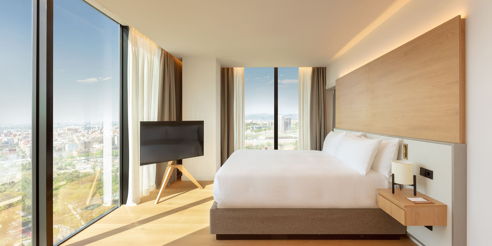 Modern hotel bedroom with king bed, panoramic city view, and wood accents.