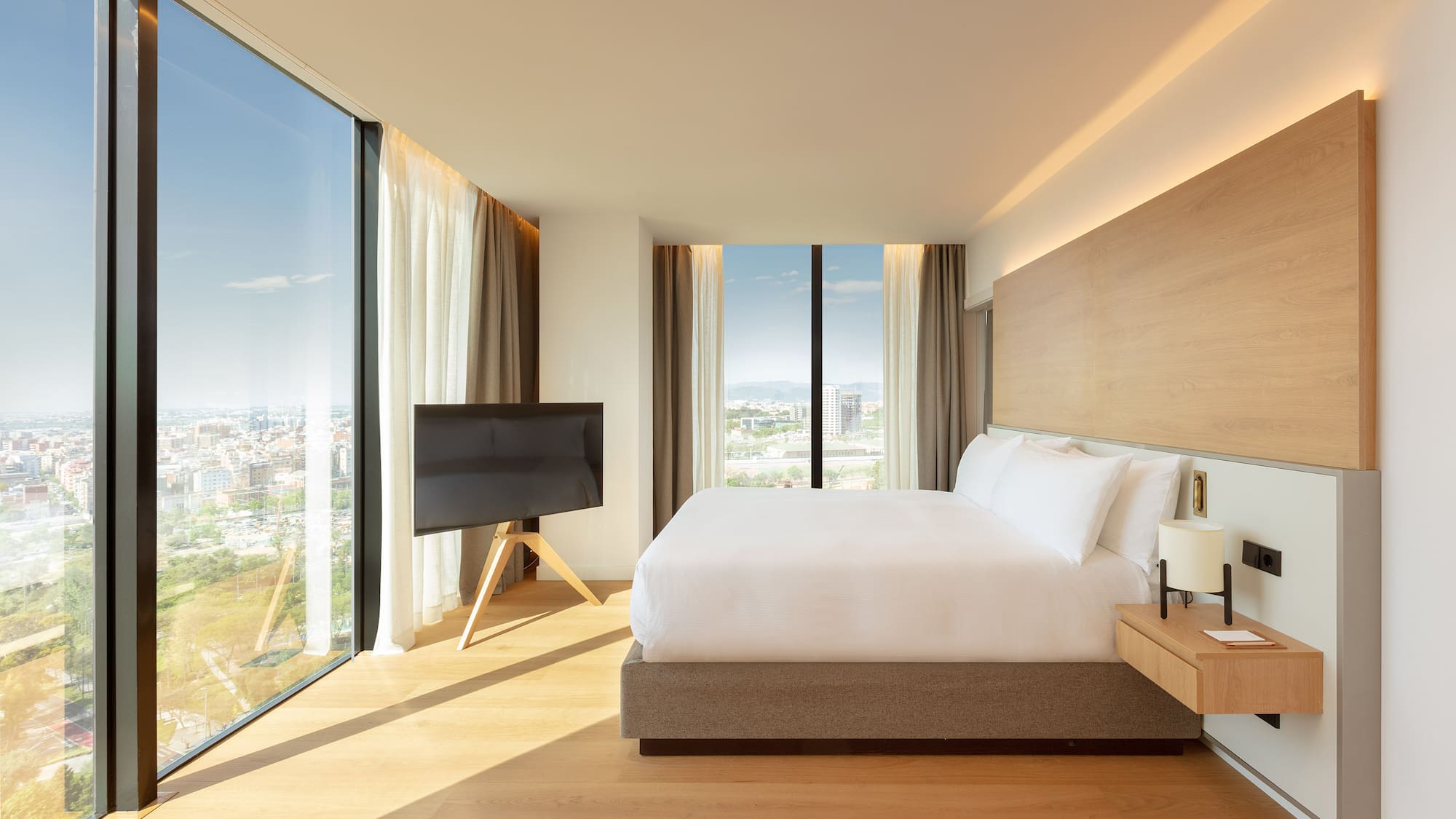 Modern hotel bedroom with king bed, panoramic city view, and wood accents.