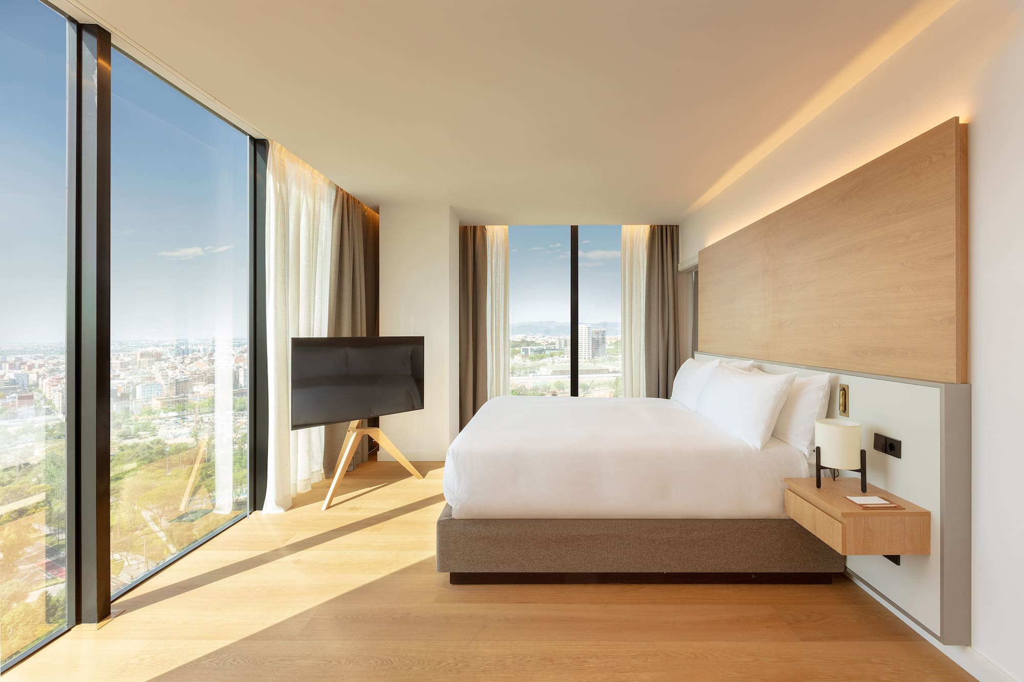 Modern hotel bedroom with king bed, panoramic city view, and wood accents.