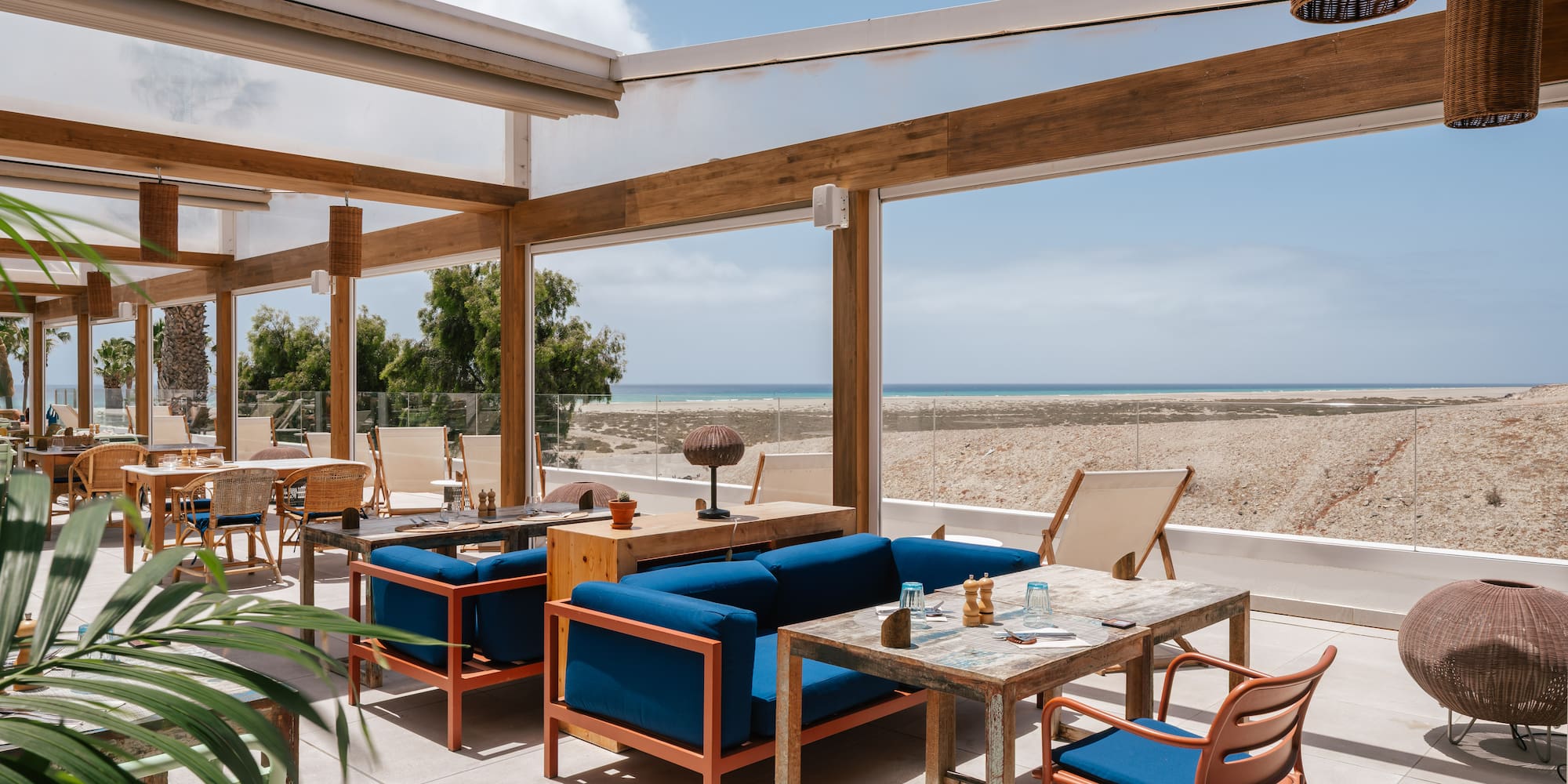 Outdoor restaurant terrace with blue seating, wooden tables, and ocean view.