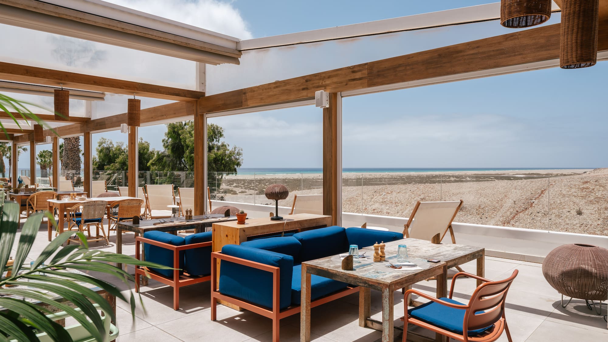 Sunny outdoor dining terrace with blue sofas, wooden tables, and ocean view.