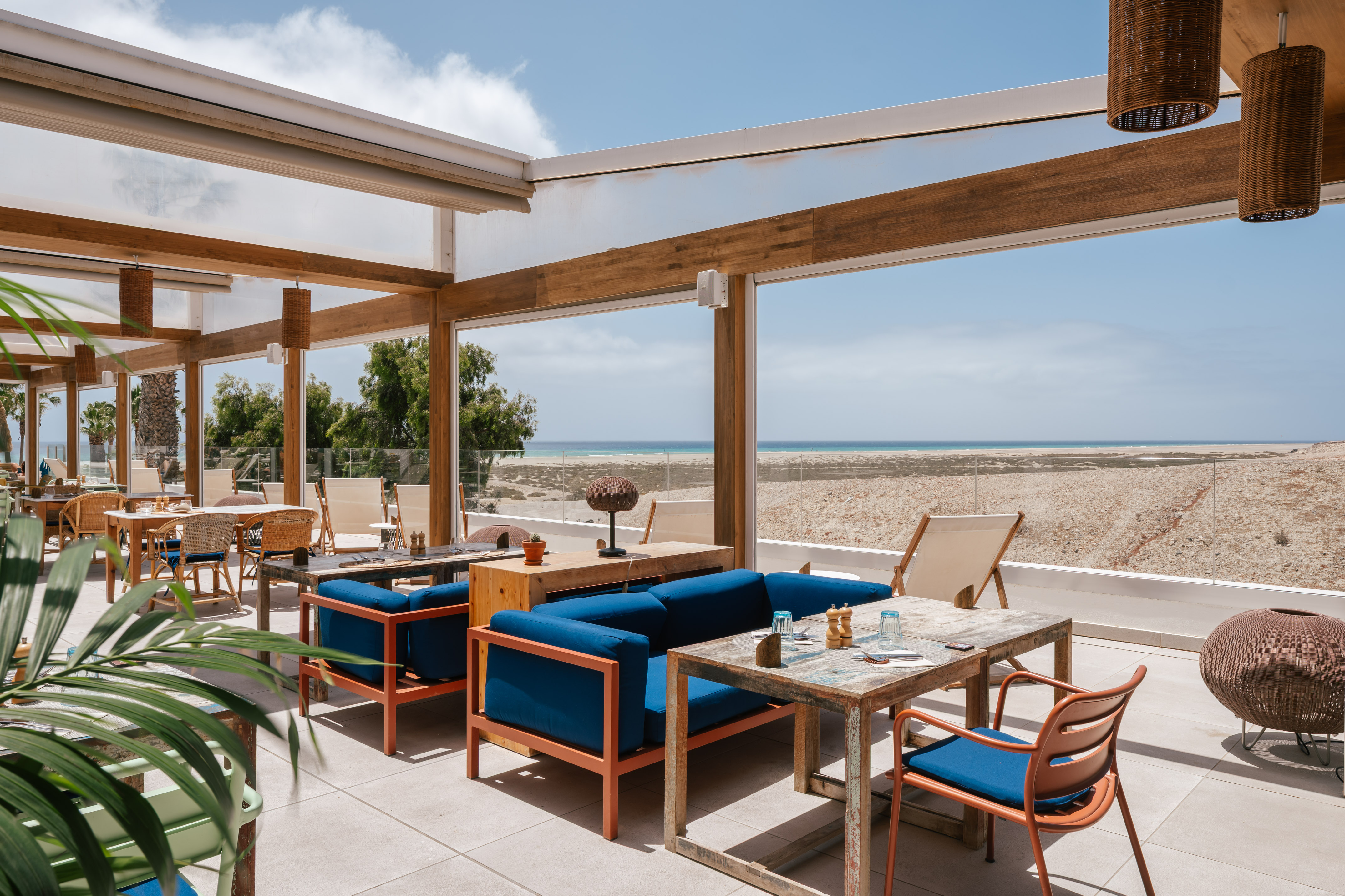 Sunny outdoor dining terrace with blue sofas, wooden tables, and ocean view.
