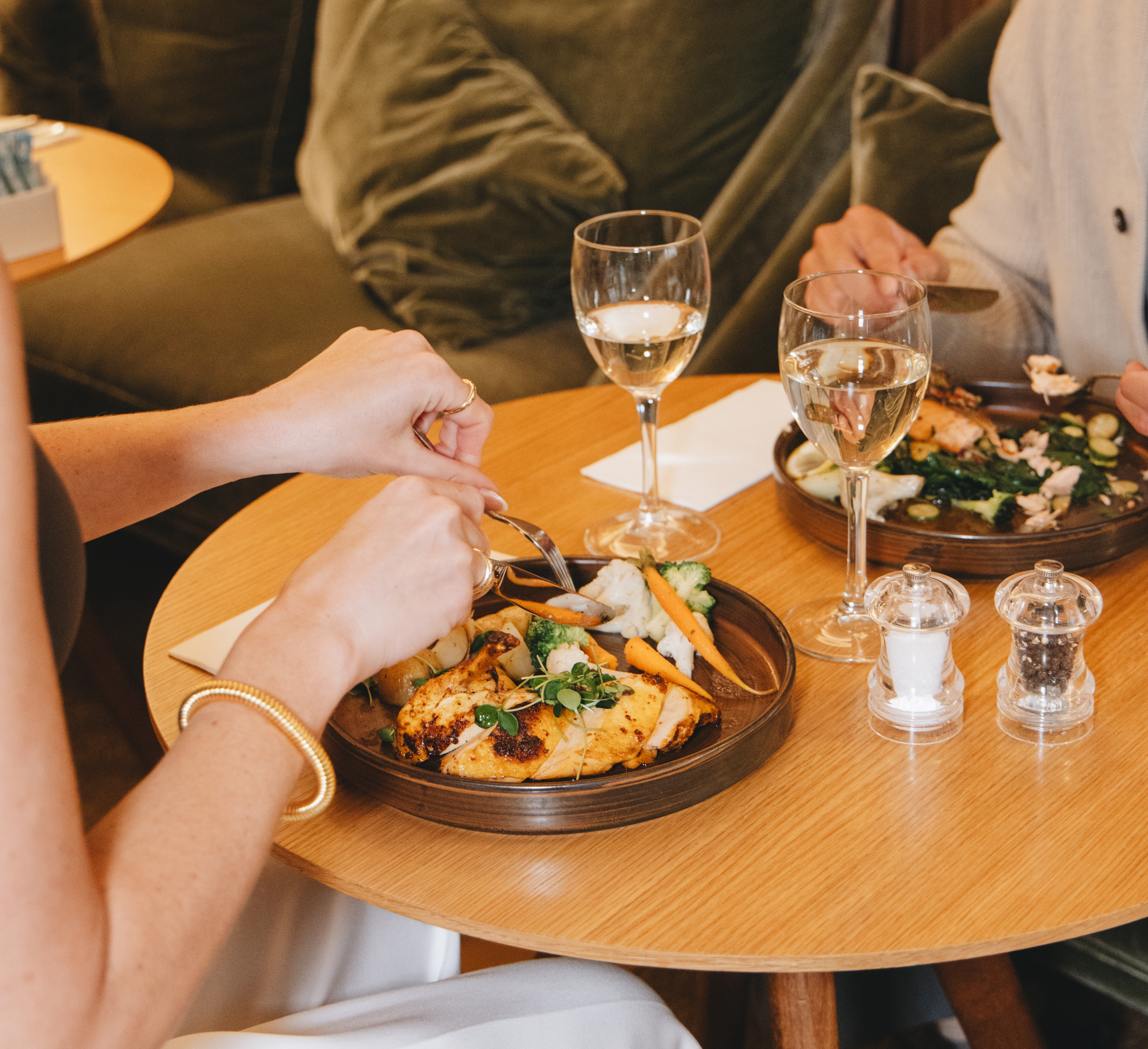 Diners enjoy roasted chicken, vegetables, and white wine at a restaurant table.