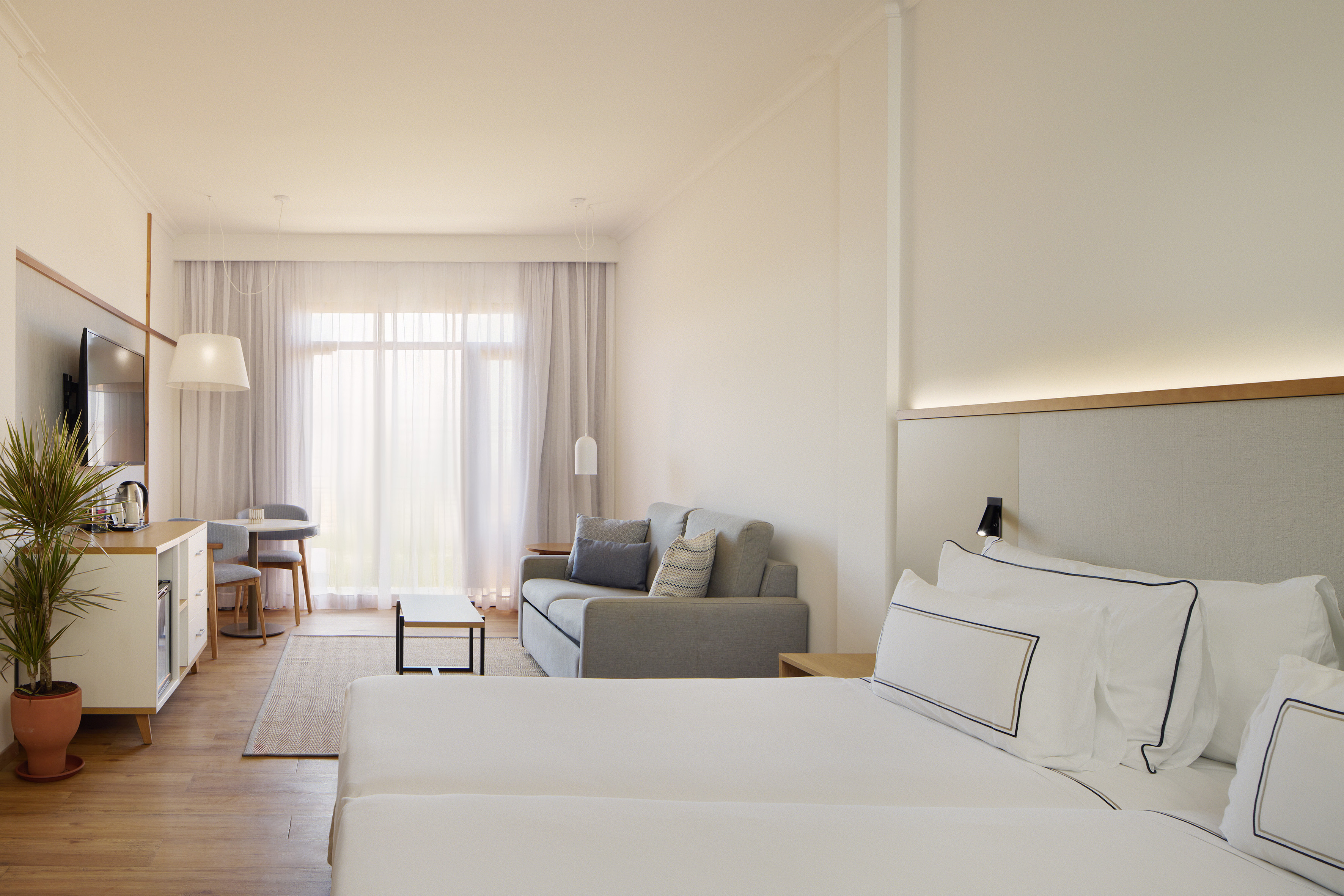 Bright modern hotel suite with bed, sofa, dining area, and large window.