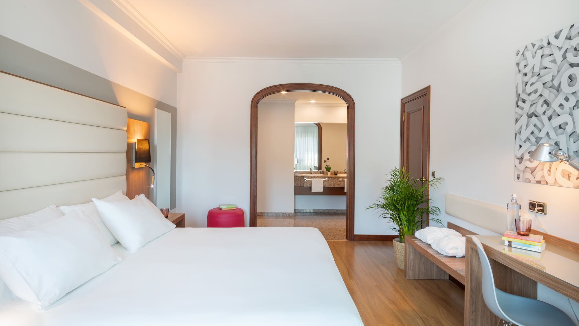 Modern hotel room with white bed, wooden desk, and arched doorway to bathroom.