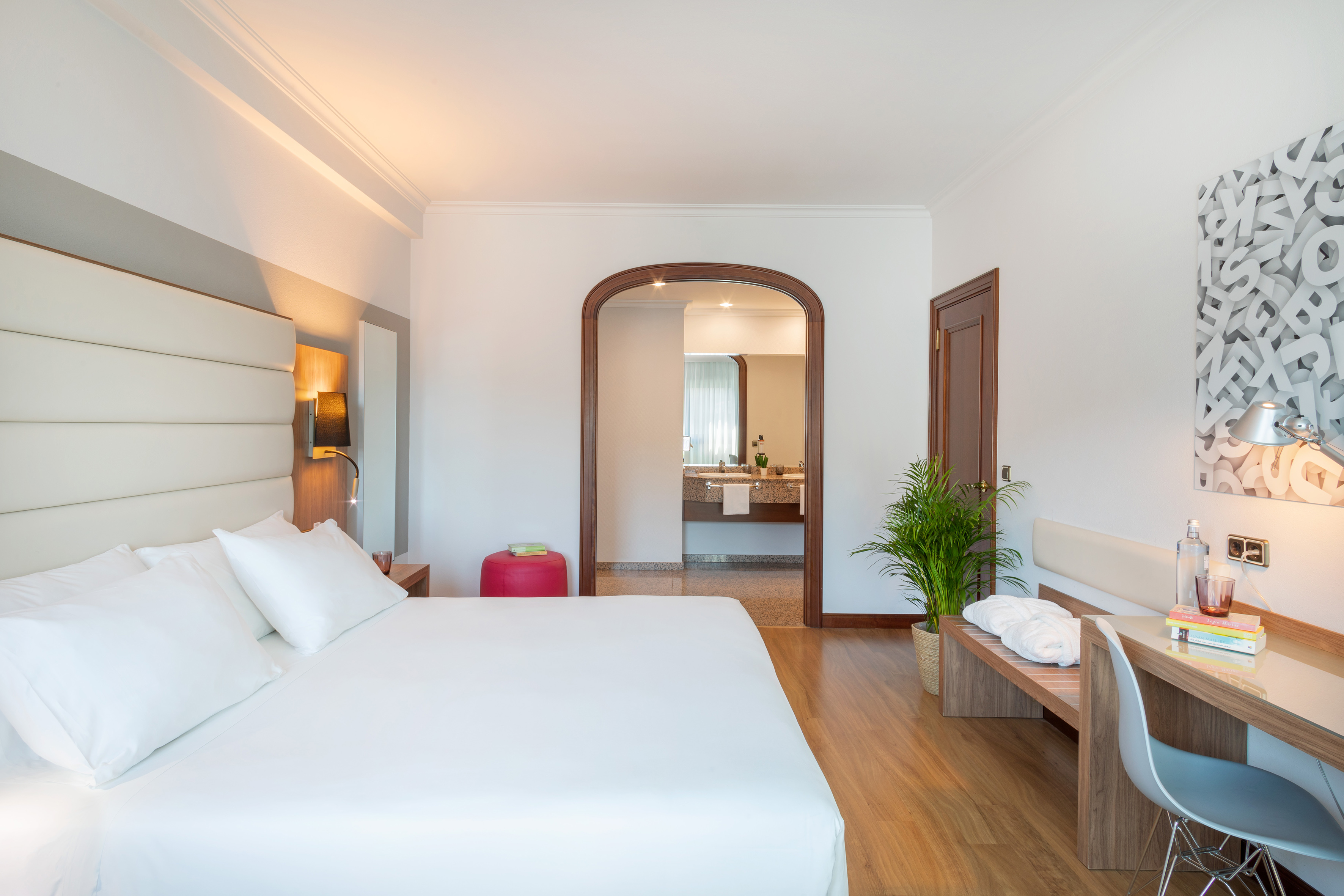 Modern hotel room with white bed, wooden desk, and arched doorway to bathroom.