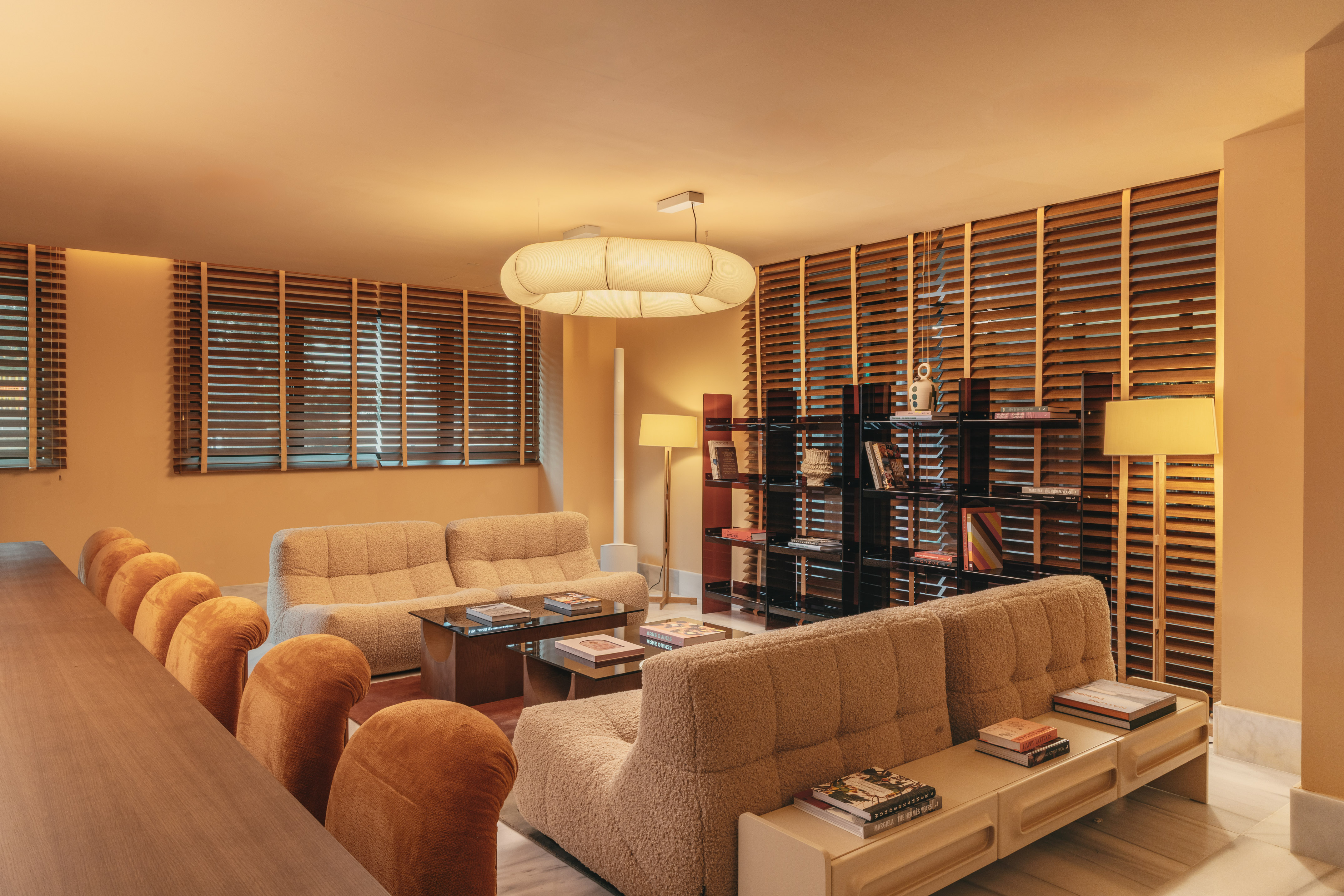 Warm retro living room with boucle sofas, modern bookshelf, and wooden slat blinds.