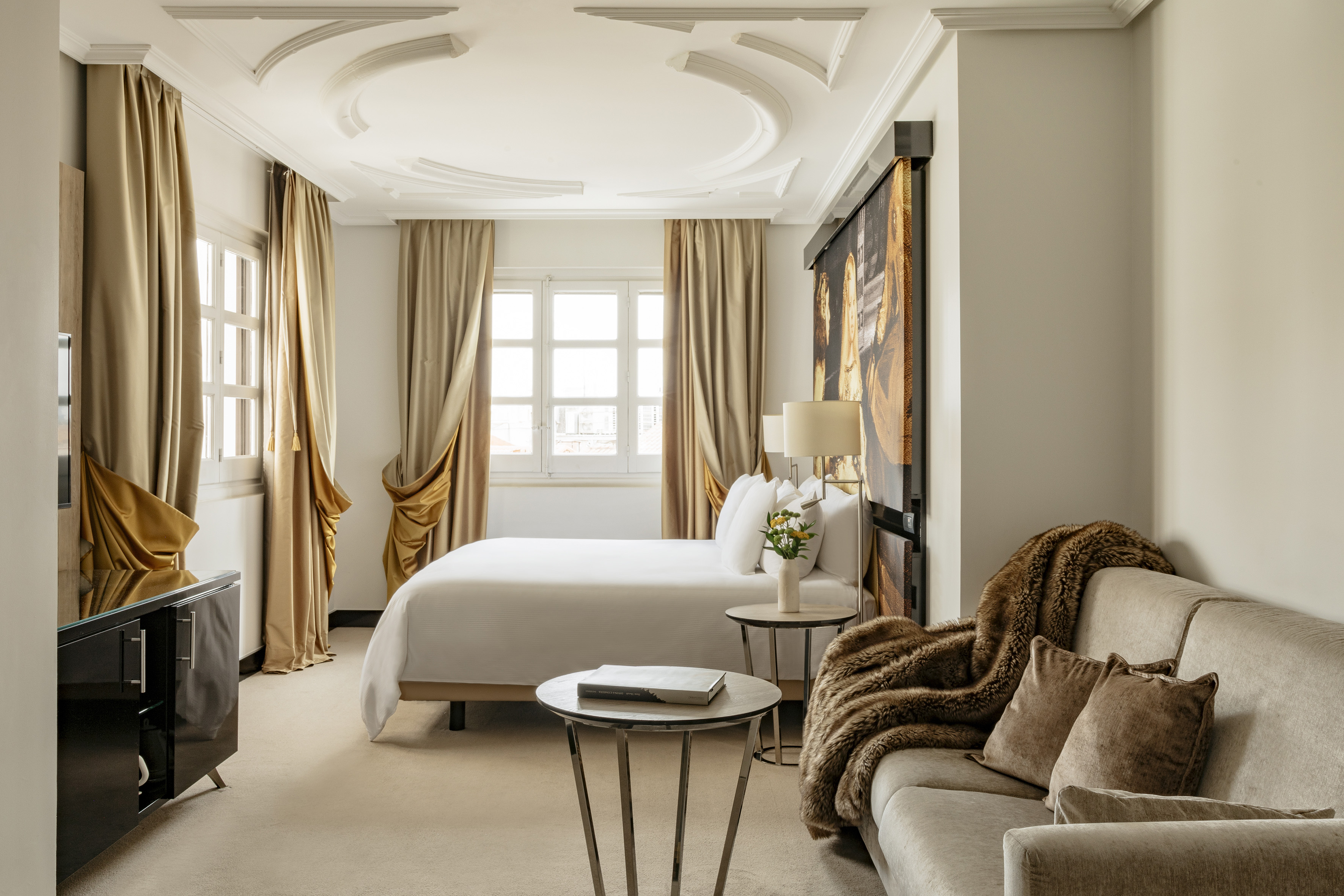 Elegant hotel room with king bed, sofa, large windows, and ornate ceiling.