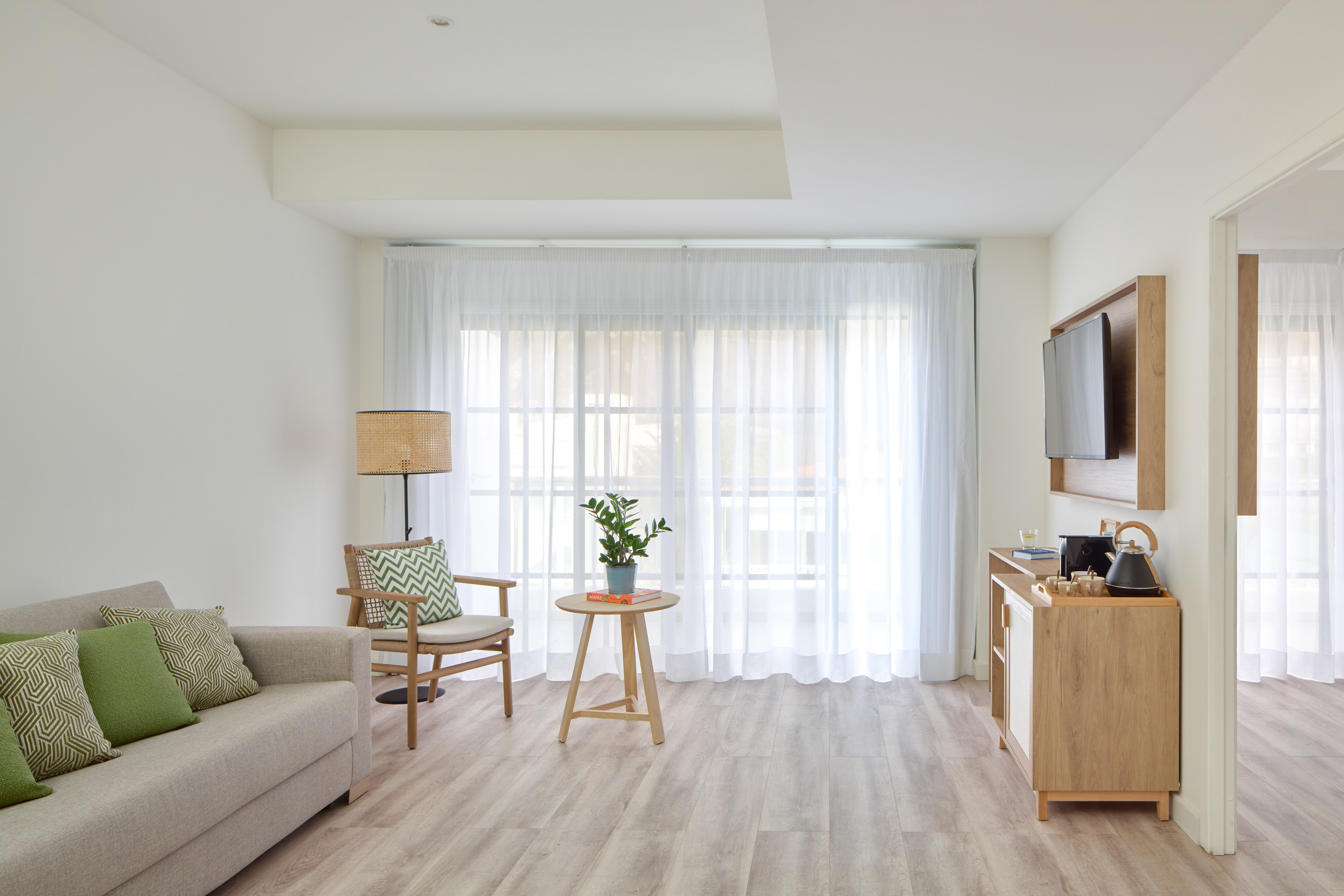 Modern hotel living room with sofa, wood furniture, sheer curtains, mounted TV.