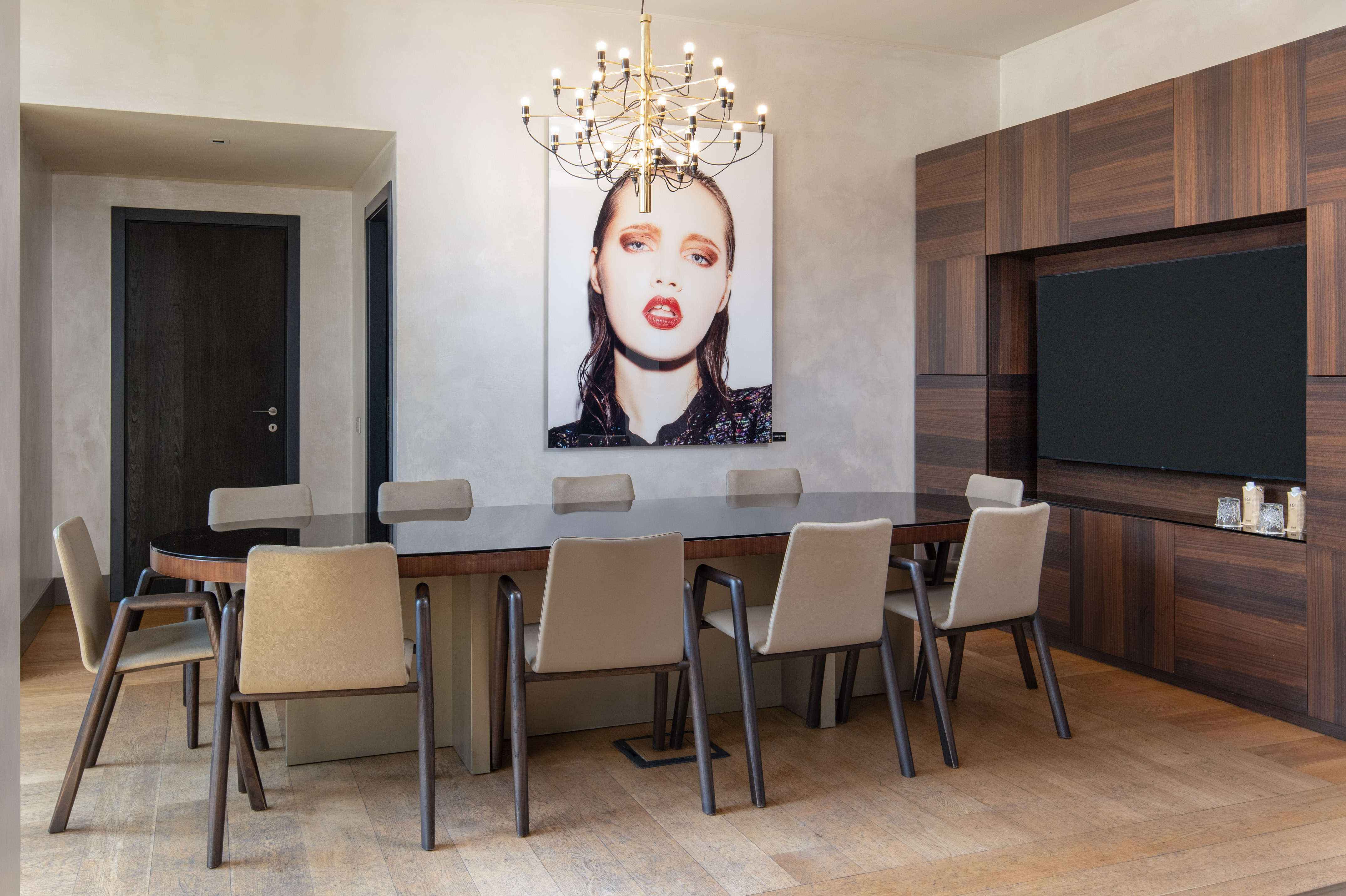 Modern dining room with oval table, beige chairs, gold chandelier, artwork, and built-in TV.