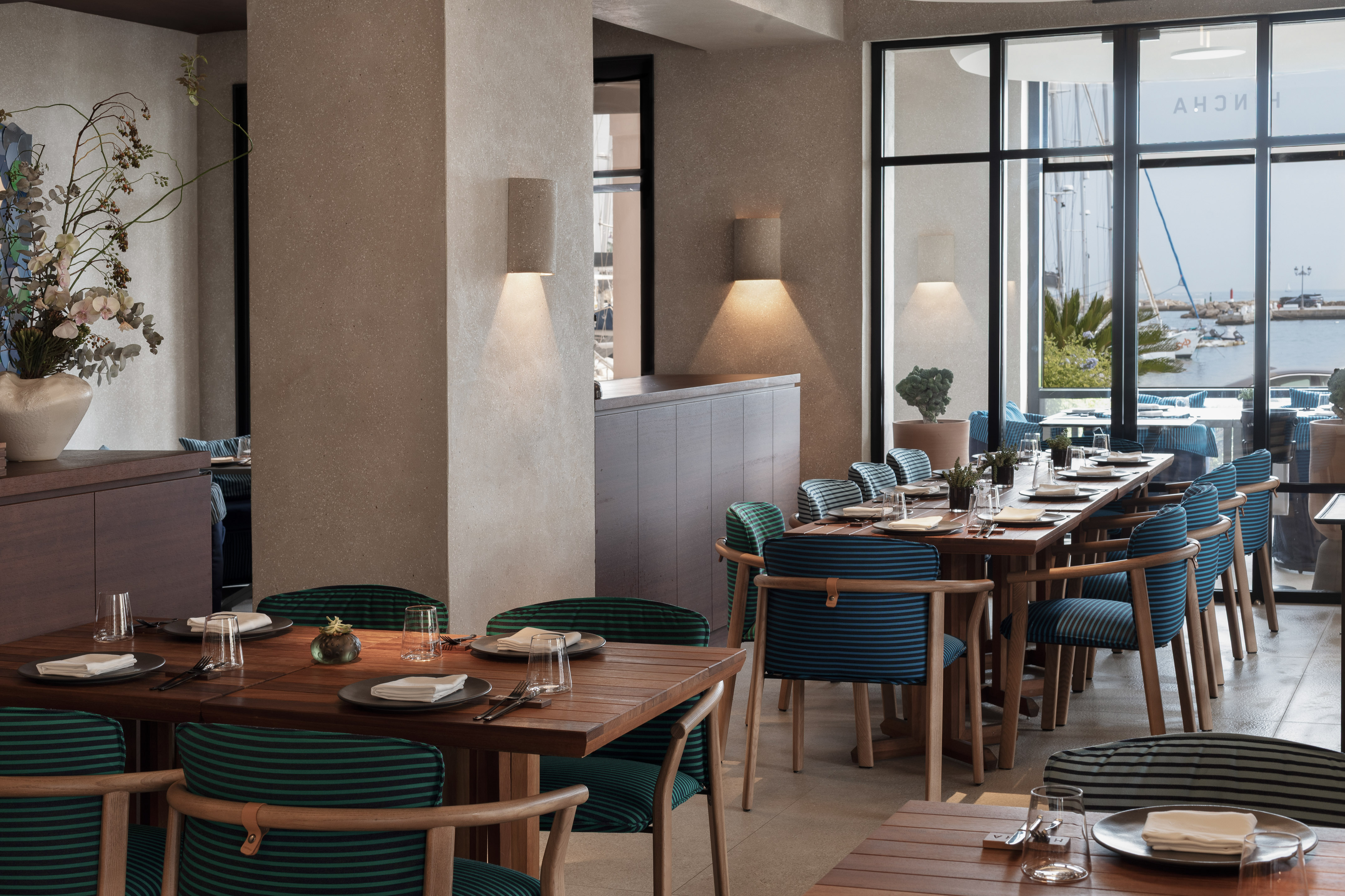 Modern restaurant interior with marina view, wooden tables, striped chairs.
