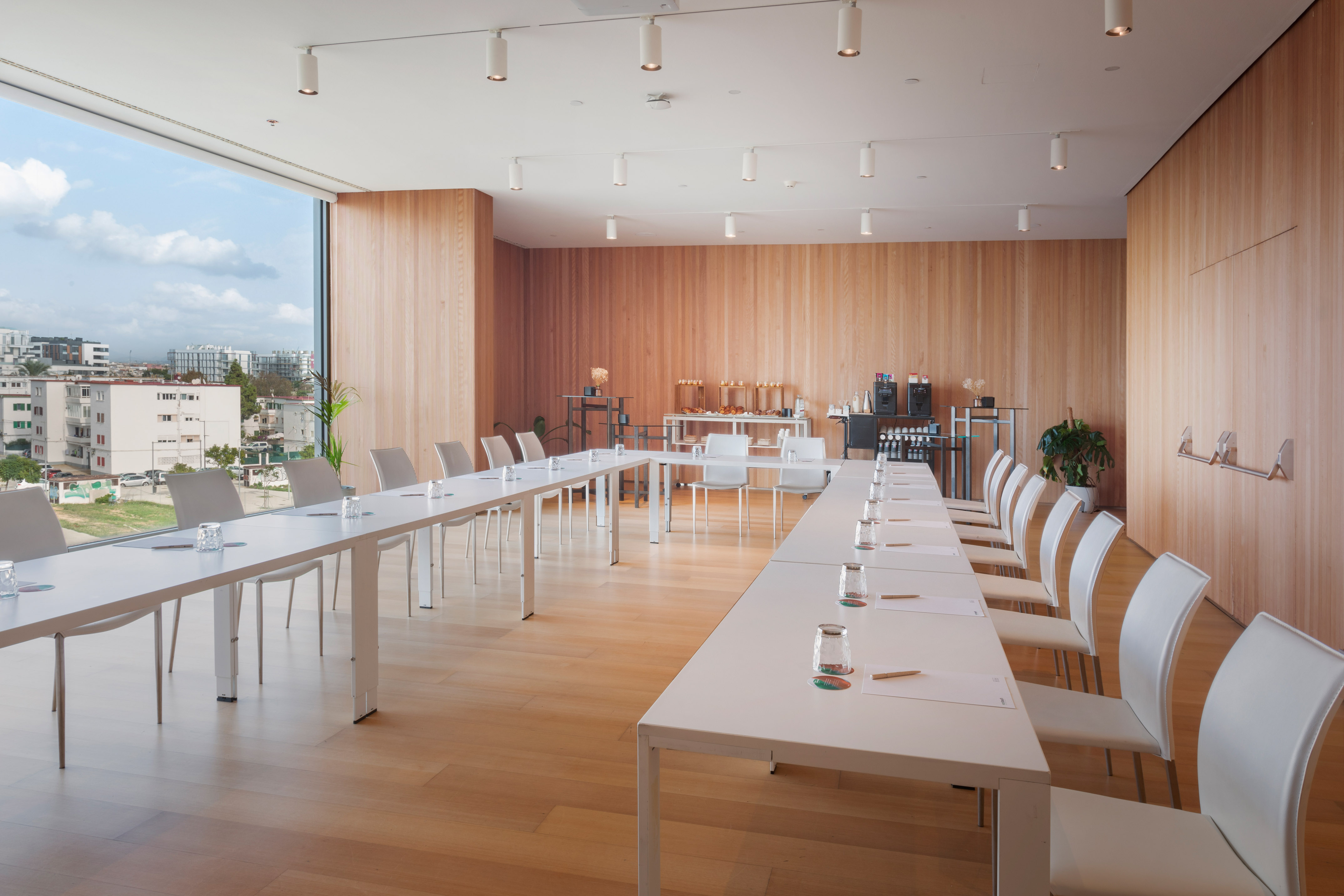 Modern meeting room with city view, wood paneling, U-shaped white tables.