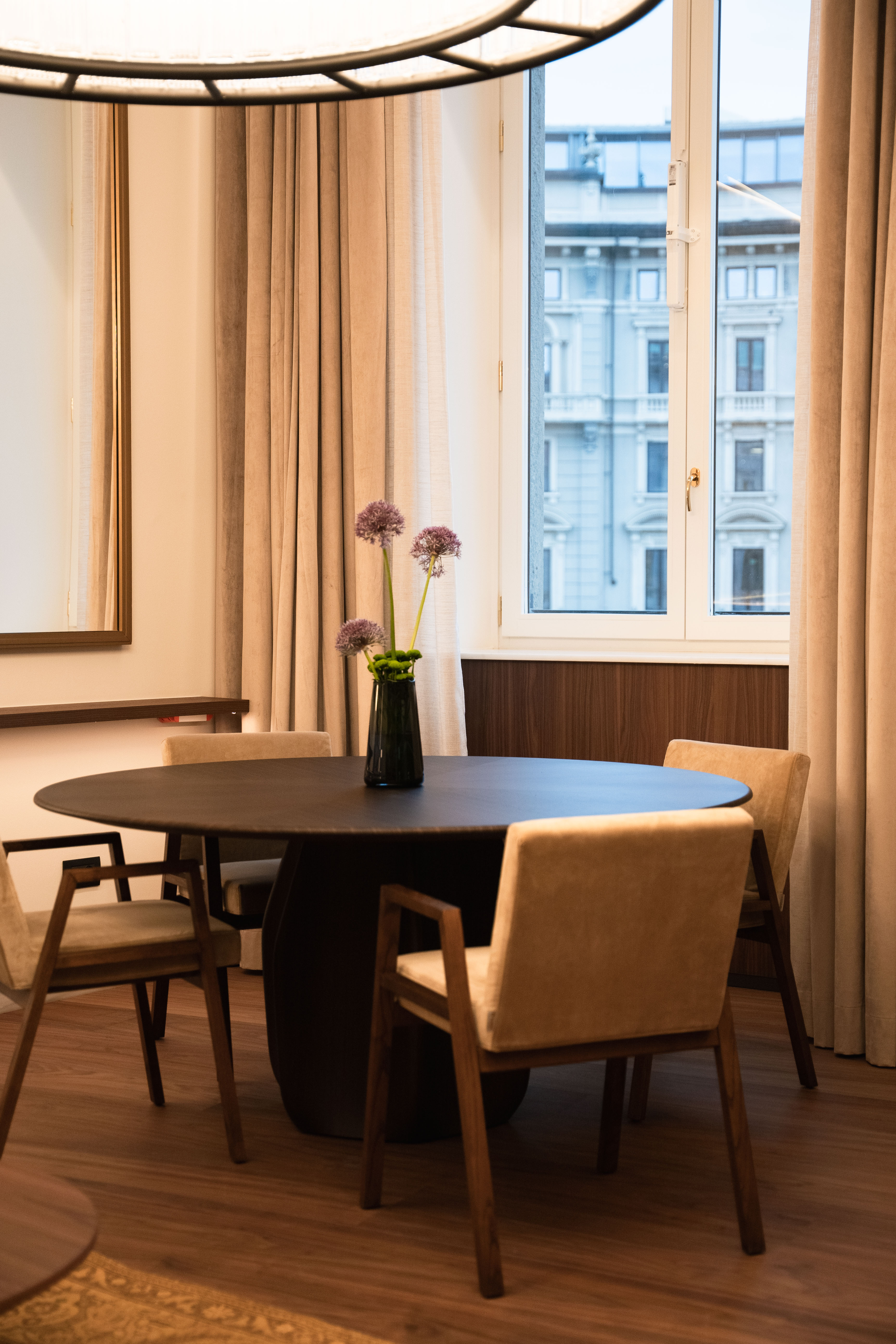 Modern dining room featuring dark round table, beige chairs, flowers, window.