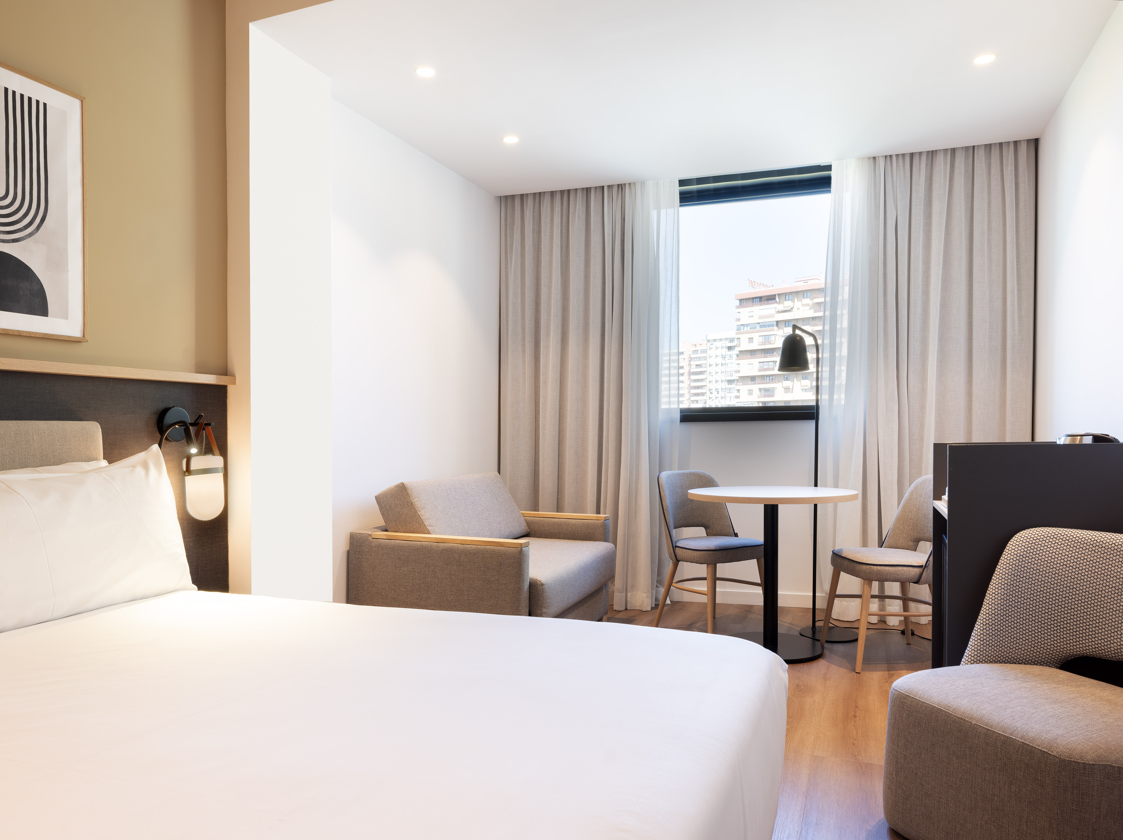 Bright modern hotel room with bed, armchair, table, and city view window.