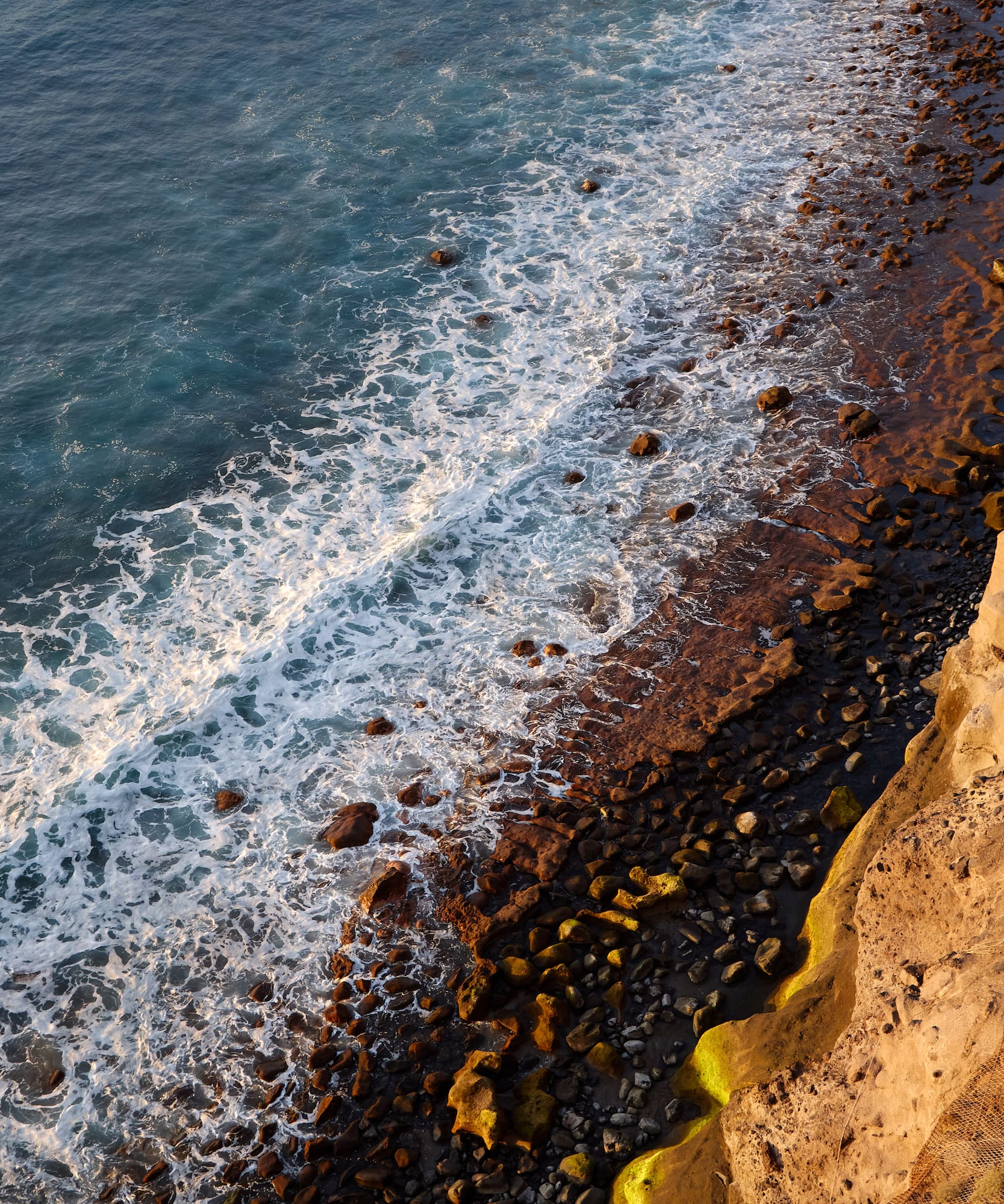 Aerial view of foamy ocean waves crashing on a rocky coastline at golden hour.