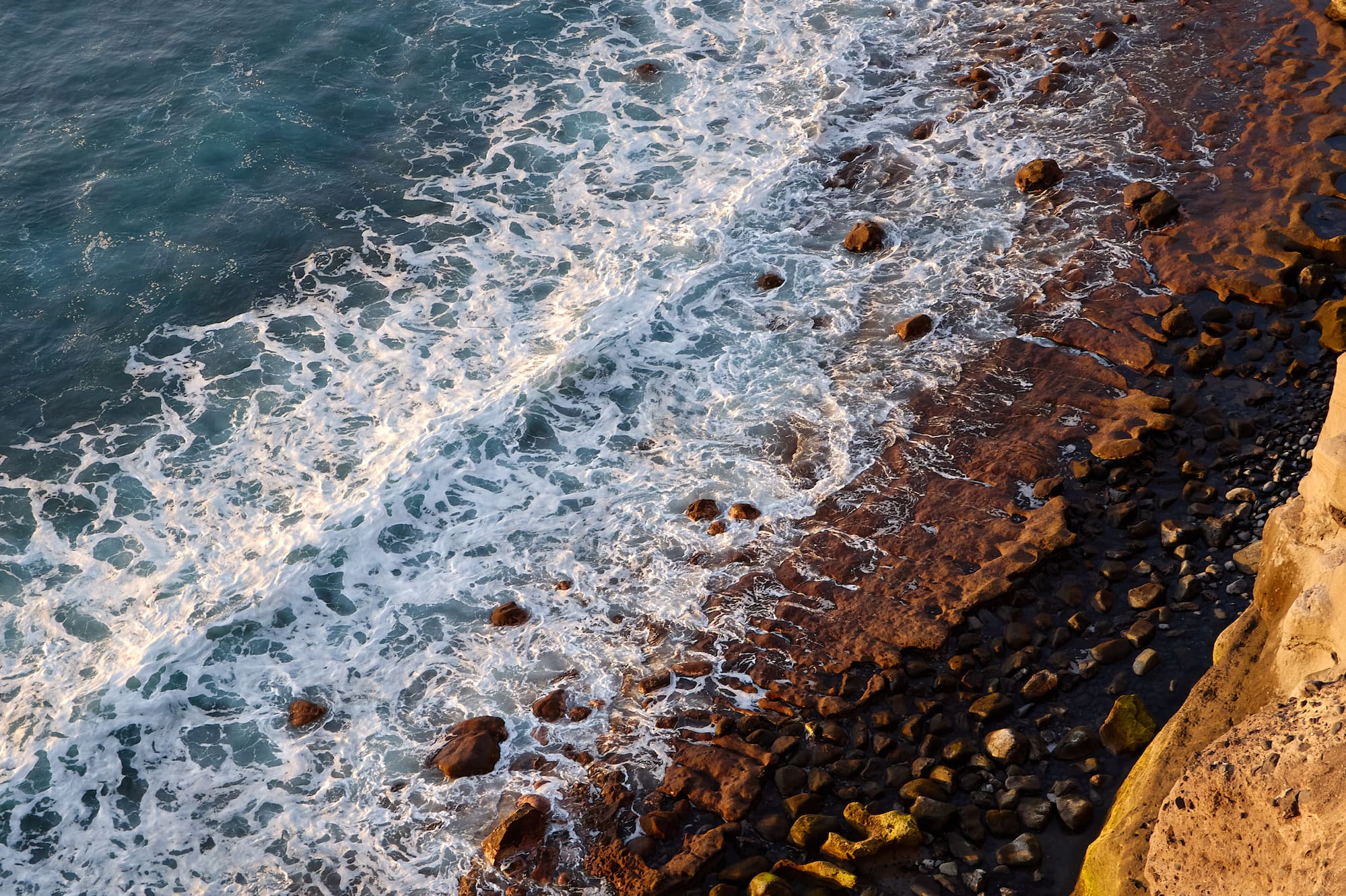 Aerial view of foamy ocean waves crashing on a rocky coastline at golden hour.