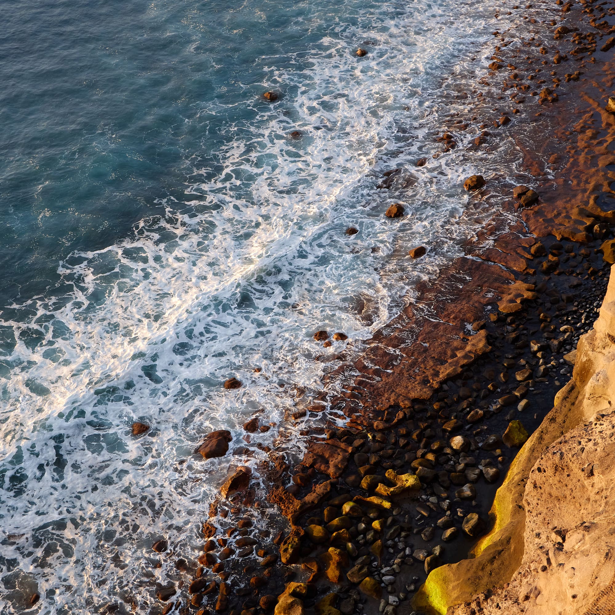 Aerial view of foamy ocean waves crashing on a rocky coastline at golden hour.