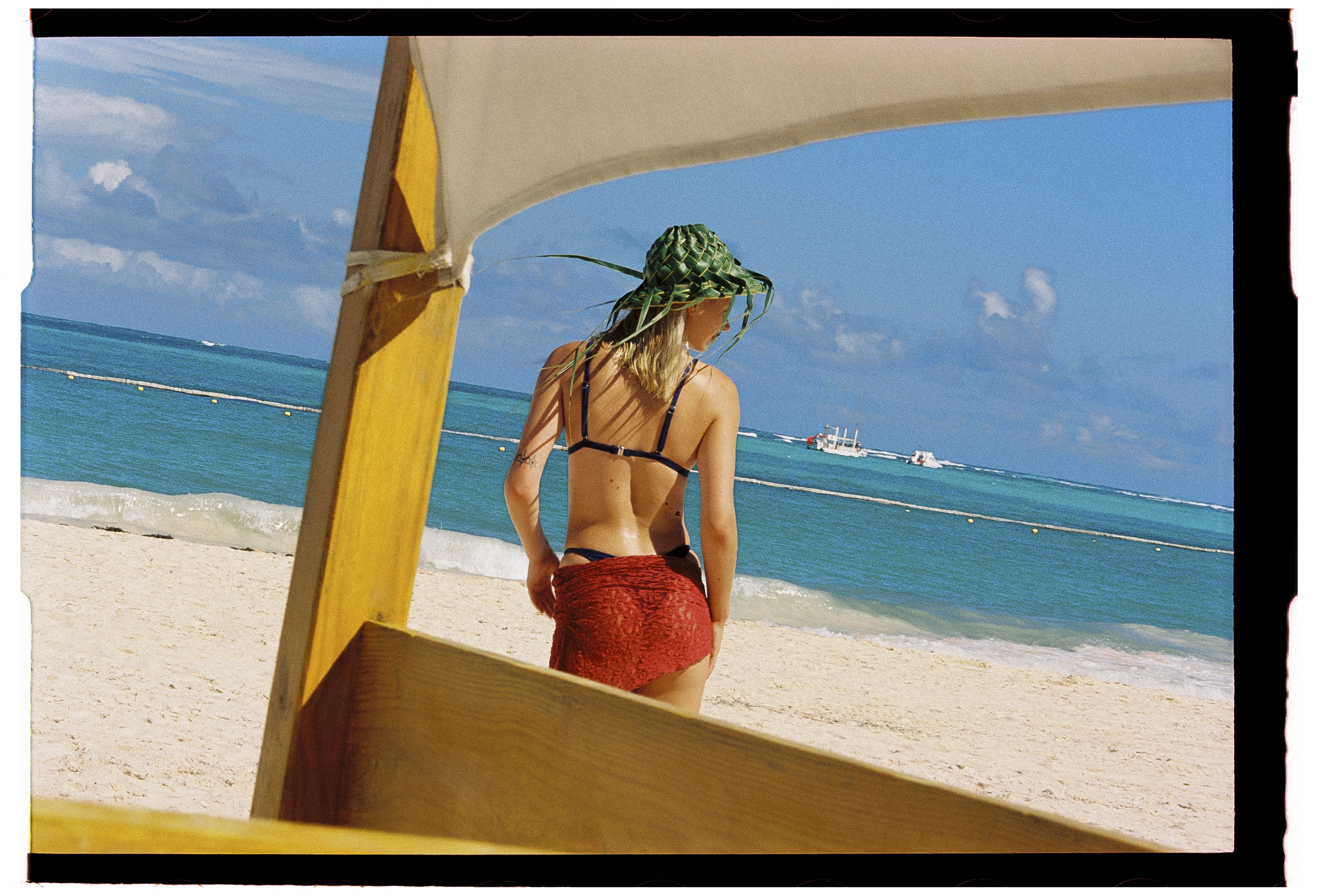 Woman in leaf hat and red sarong on beach, looking at turquoise ocean.