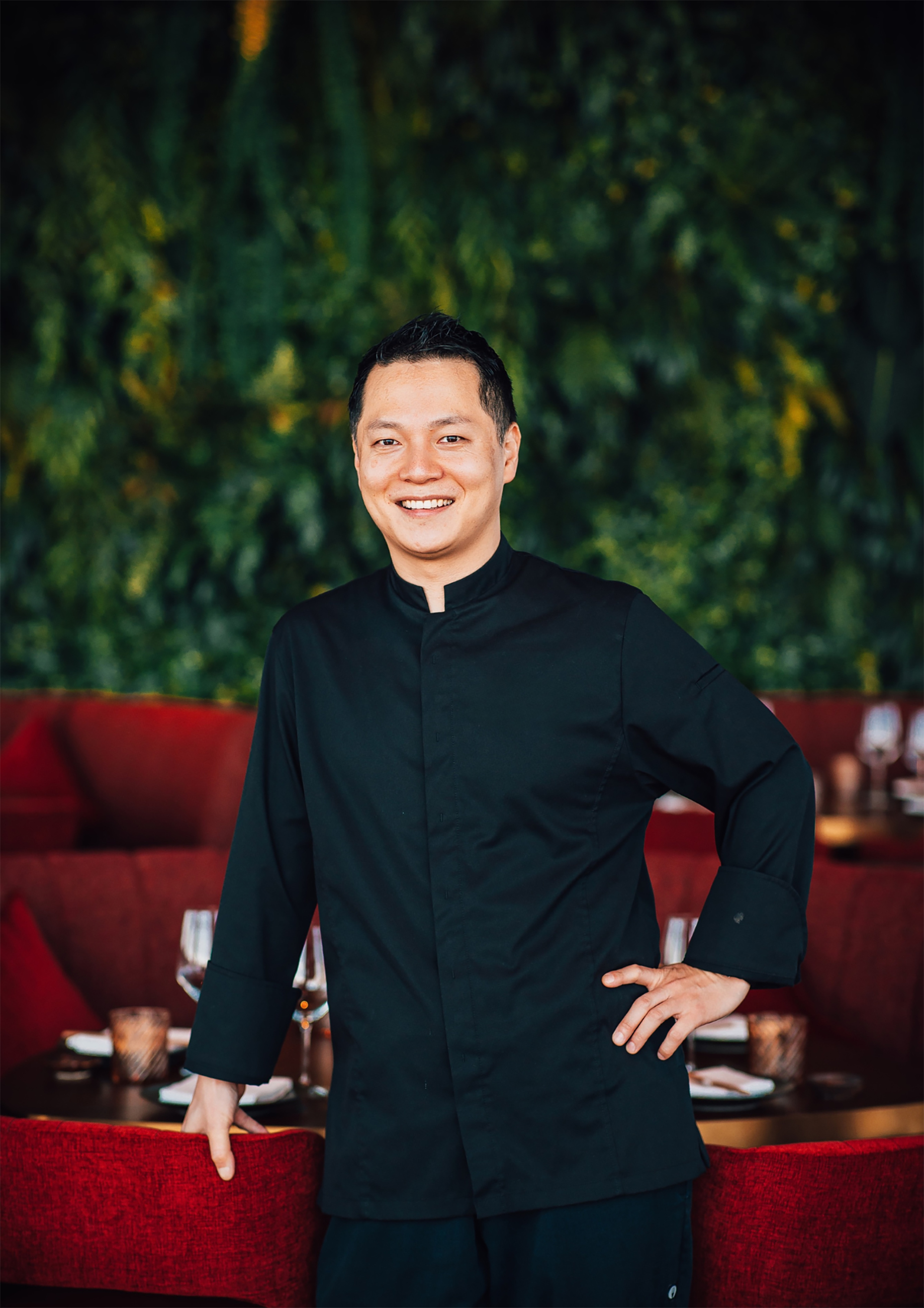 Smiling Asian chef in black uniform, restaurant with red seating & green wall.